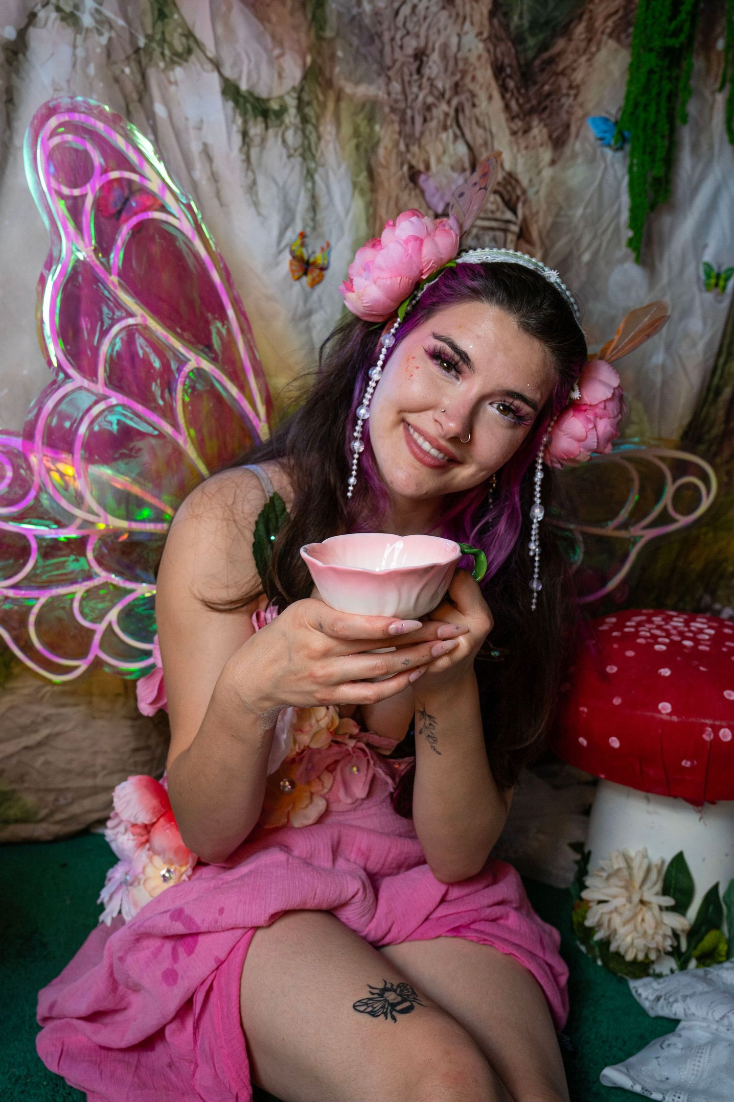 Smiling fairy costume model with pink wings holds a pink teacup beside red mushrooms in enchanted forest set. Fantasy Photoshoot