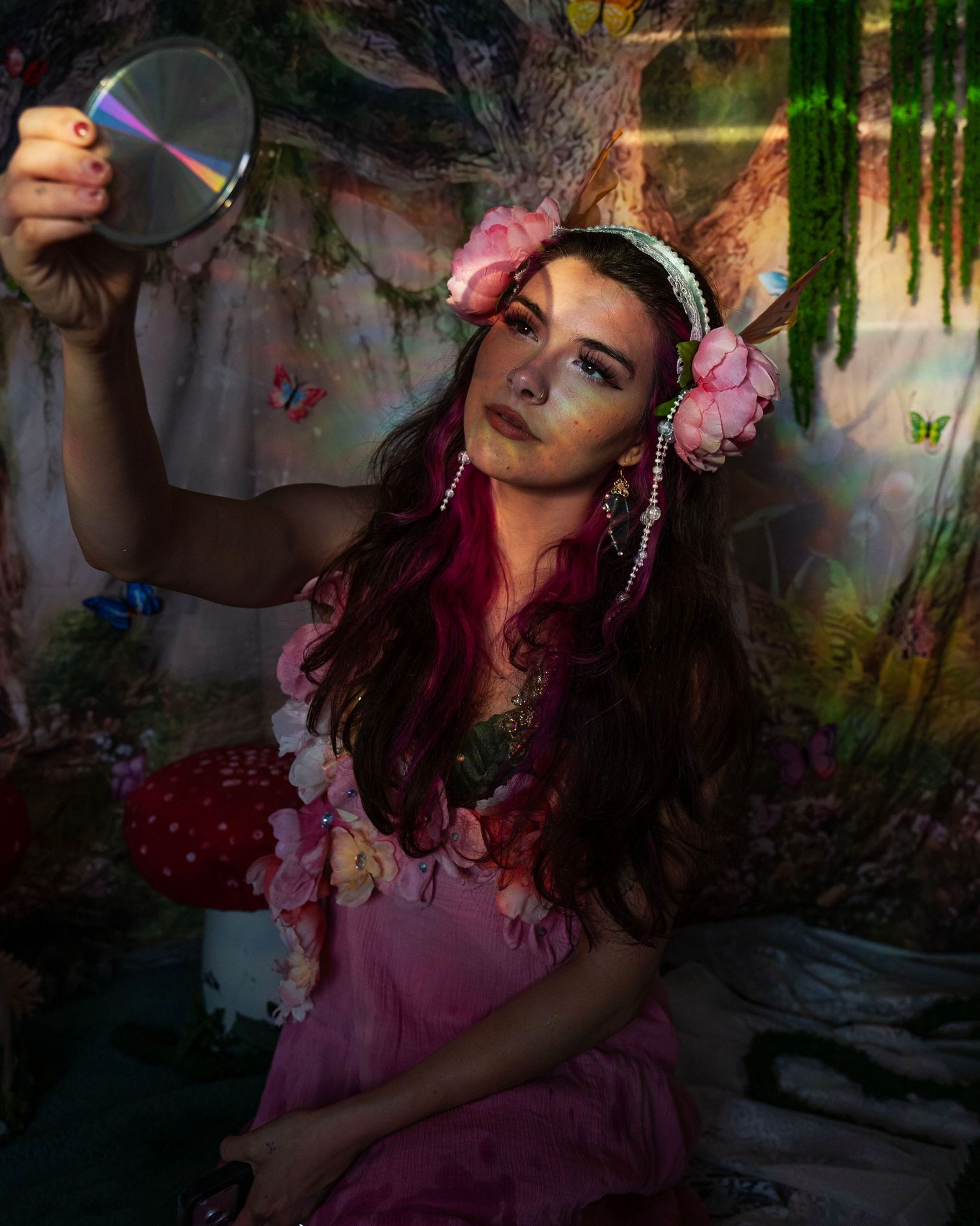 Woman in floral fairy costume holds a reflective disc mirror in a magical forest backdrop studio. Fantasy Photoshoot