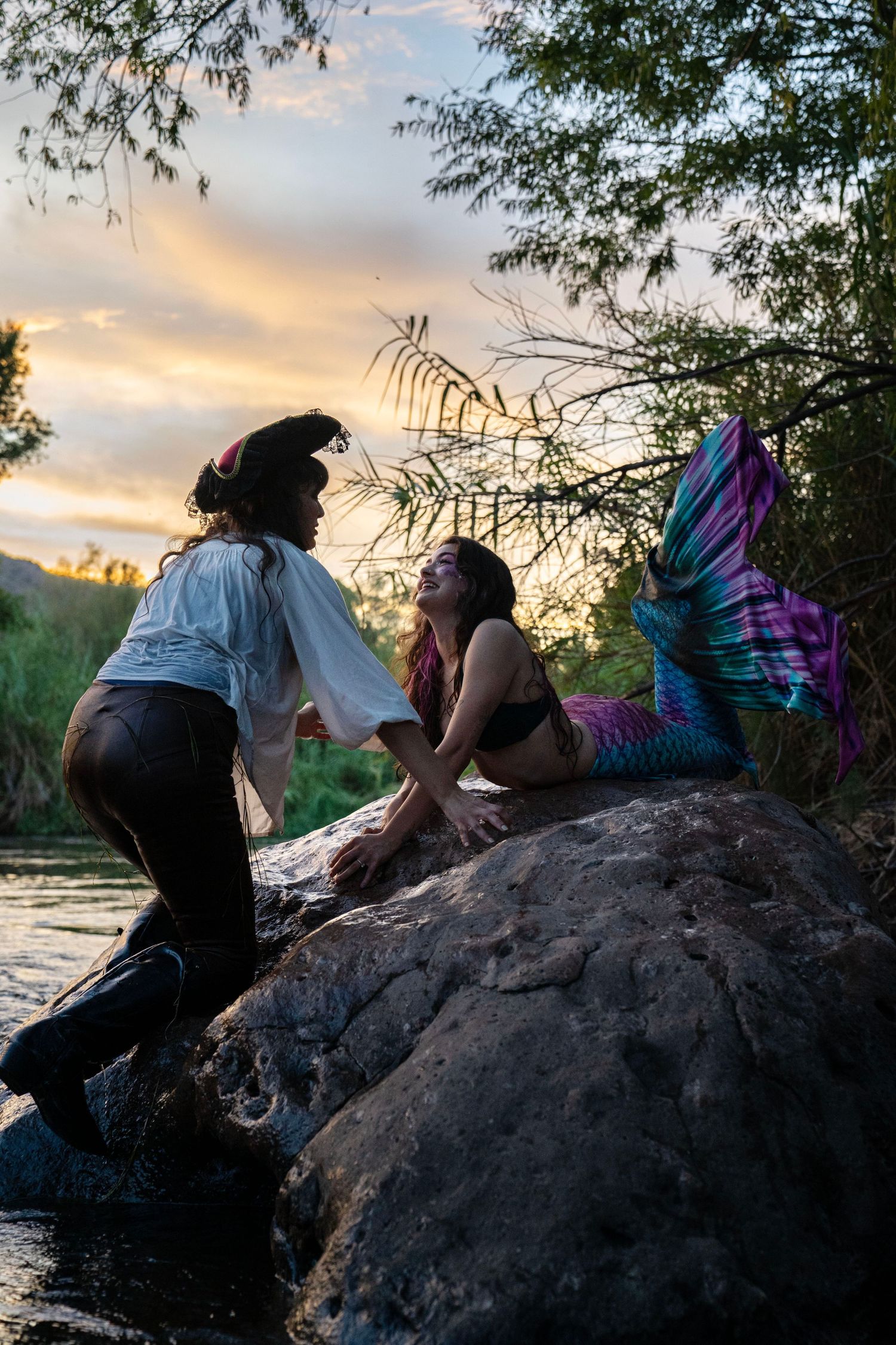 Pirate and mermaid cosplay duo posing on rocks by a river at golden hour sunset. Mermaid photoshoot, fantasy photoshoot.