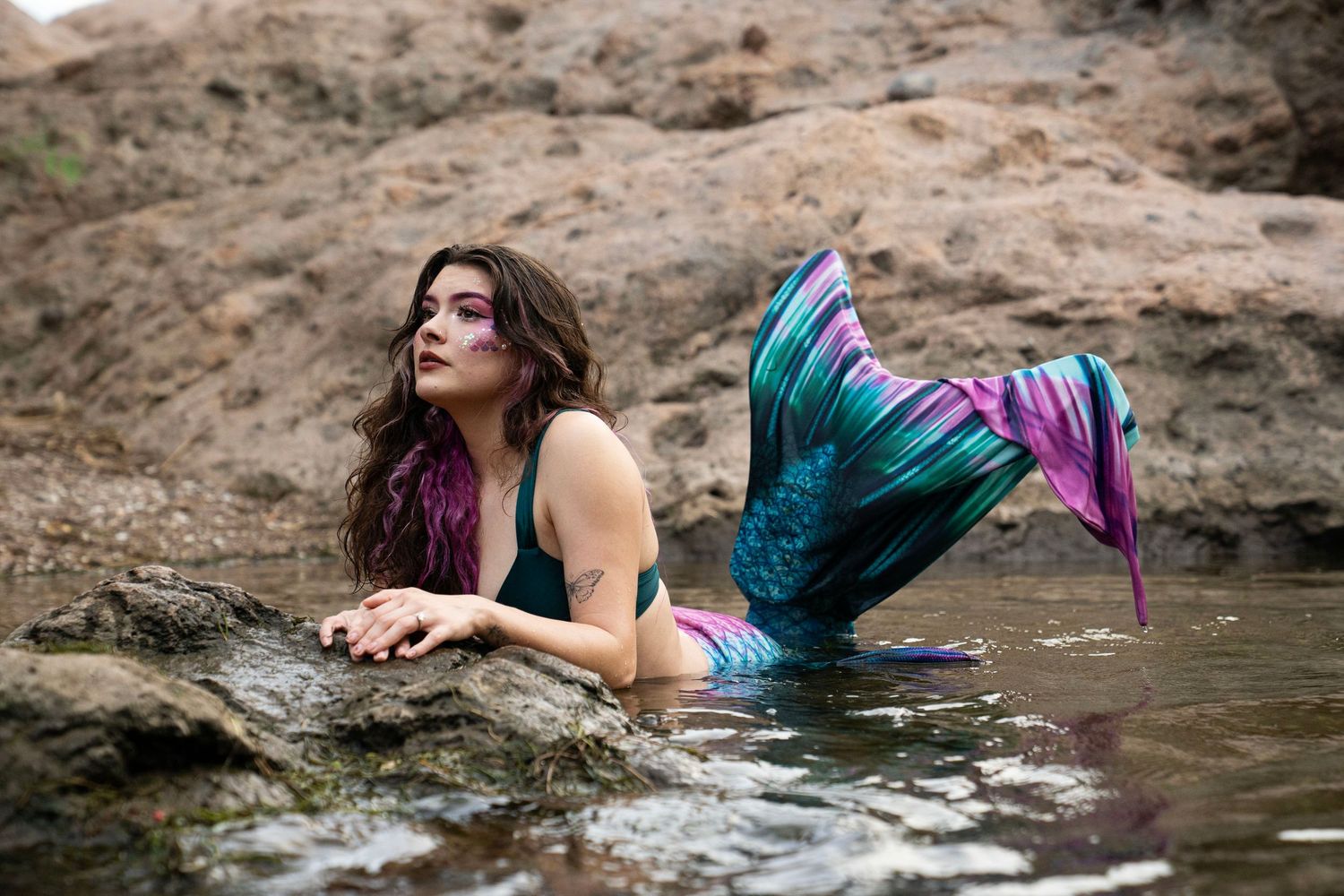 A tattooed woman in a teal mermaid costume with vibrant tail reclines on rocks near water. Fantasy Photoshoot