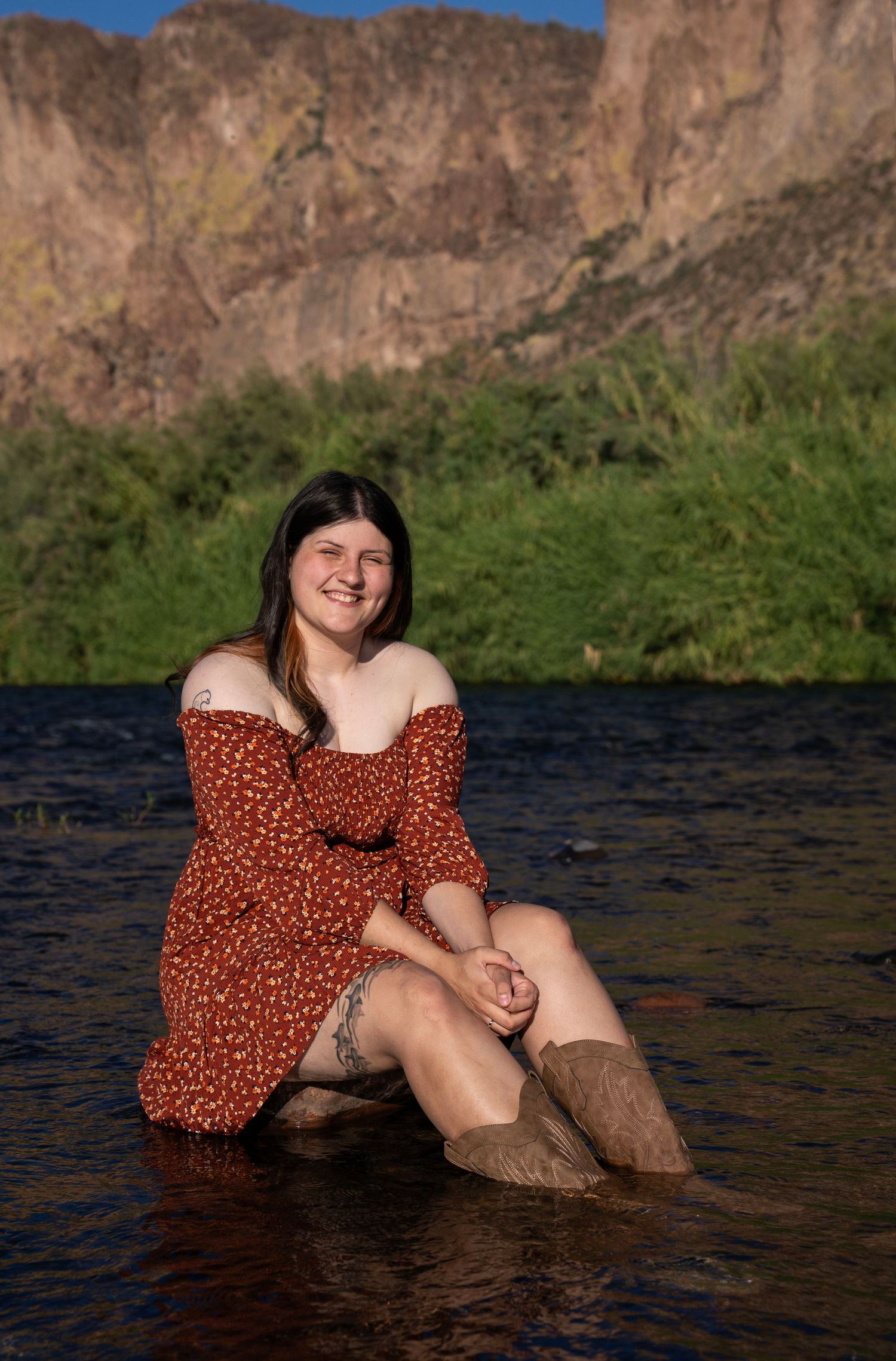 Young woman in red floral off-shoulder dress sitting in shallow river with rocky mountain backdrop.