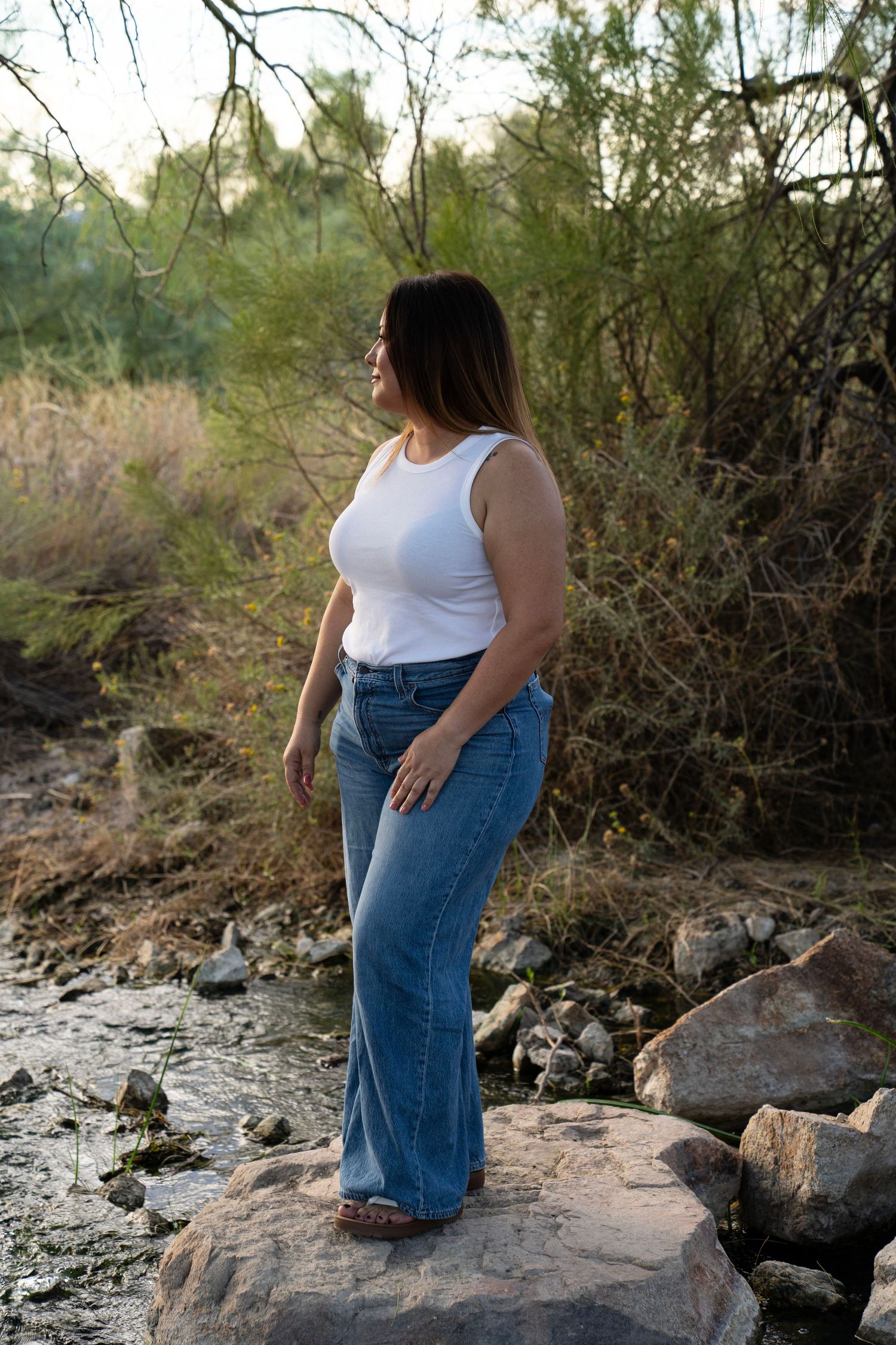 Woman in white tank top and flare jeans standing on rocks by a stream in a natural outdoor setting.