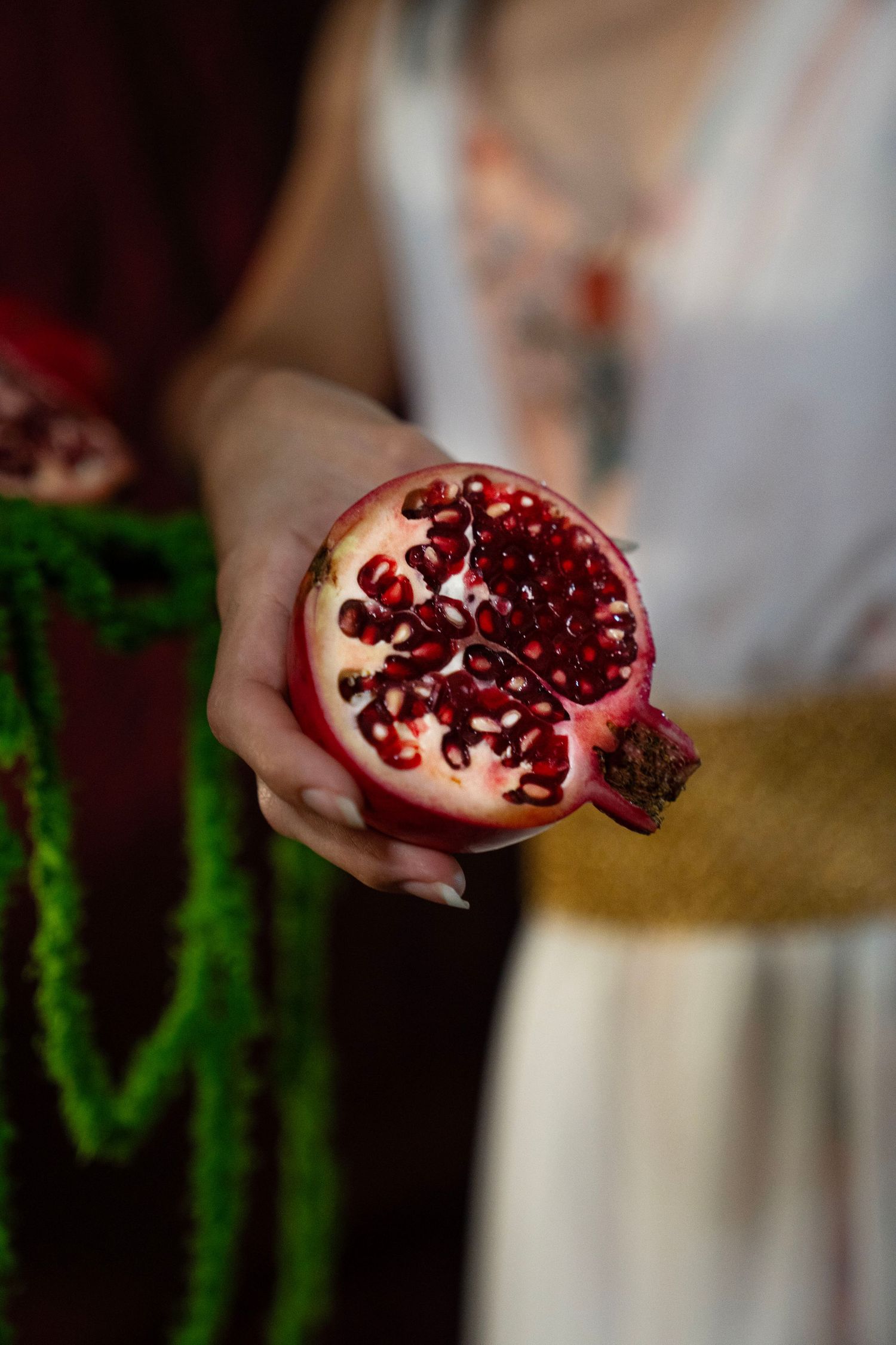 Woman in a white dress with a gold sash holds out a halved pomegranate showing ruby red seeds, with green garland in the background.