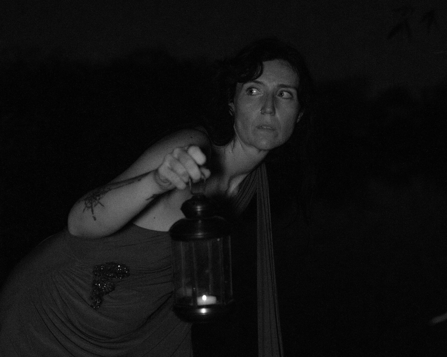 Woman with tattooed arm holds a lit candle lantern in a dark night scene, glancing sideways in this moody black and white portrait.