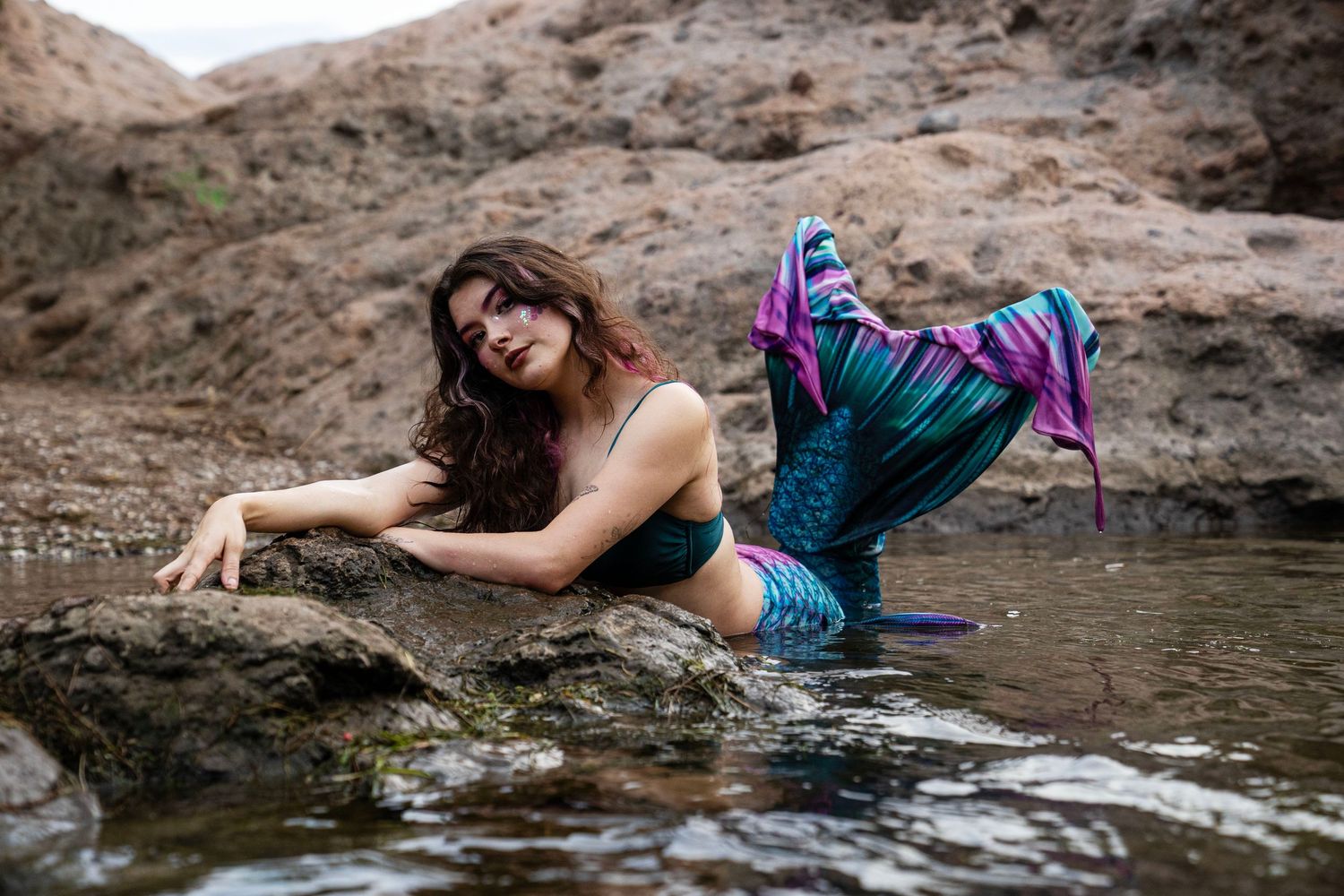 A young woman in a teal mermaid bikini top and colorful tail reclines gracefully on wet rocks. Fantasy Photoshoot