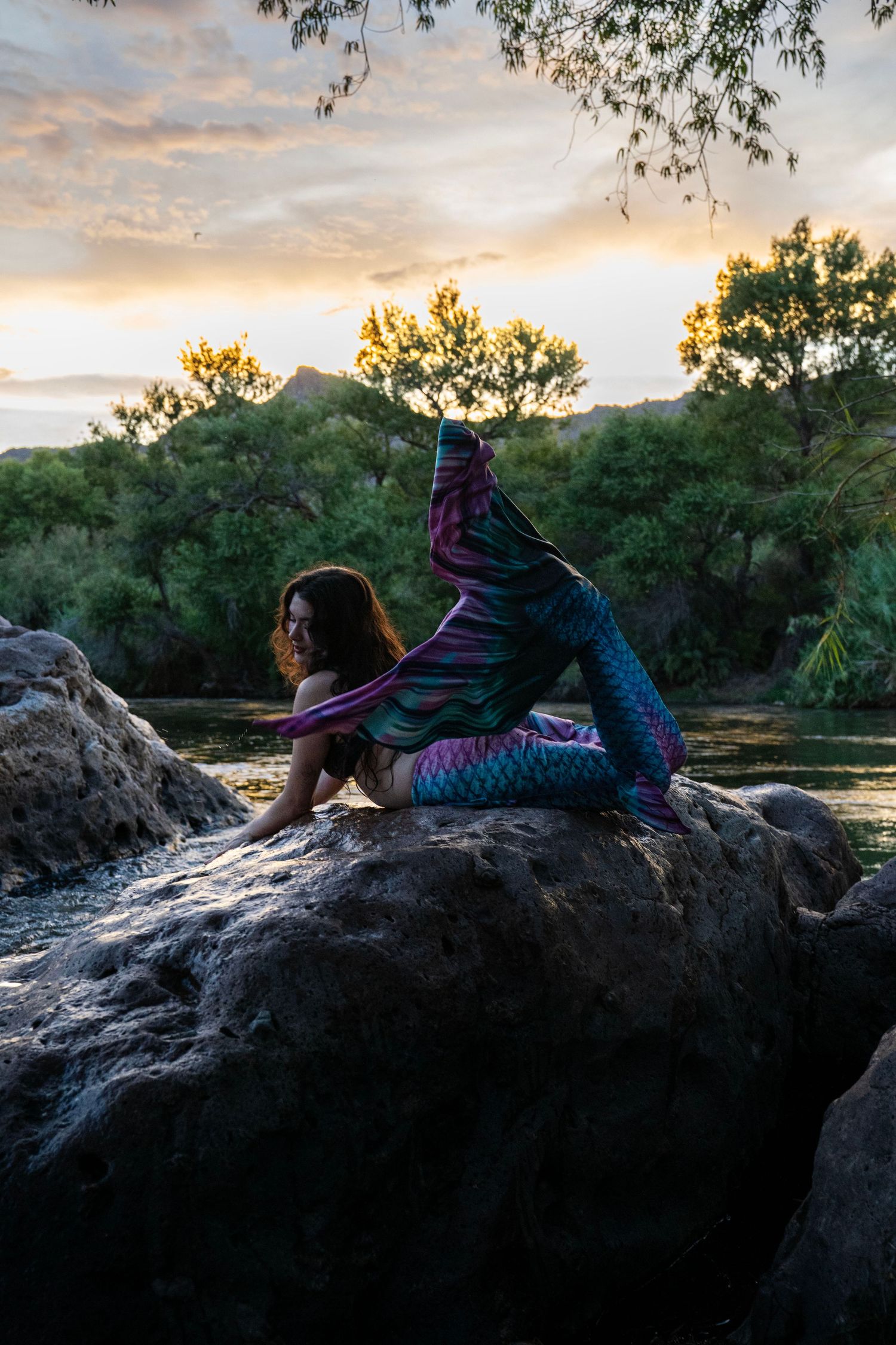 Woman in vibrant mermaid tail costume poses dramatically on rock by river at sunset. Mermaid photoshoot, fantasy photoshoot.