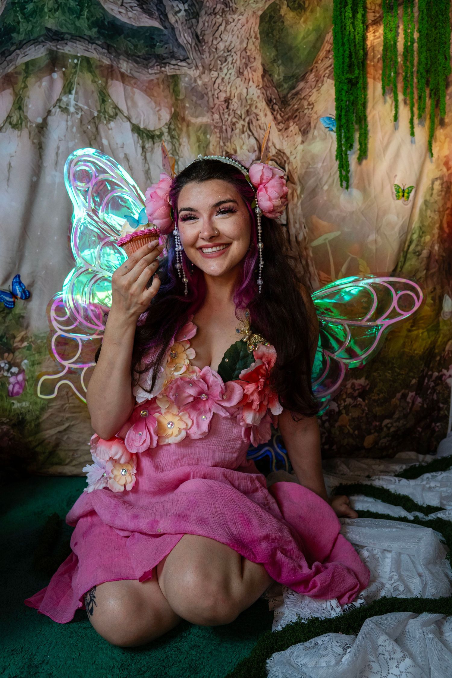 Smiling fairy with glowing wings and pink floral dress holds a cupcake in a magical enchanted forest backdrop. Fantasy Photoshoot