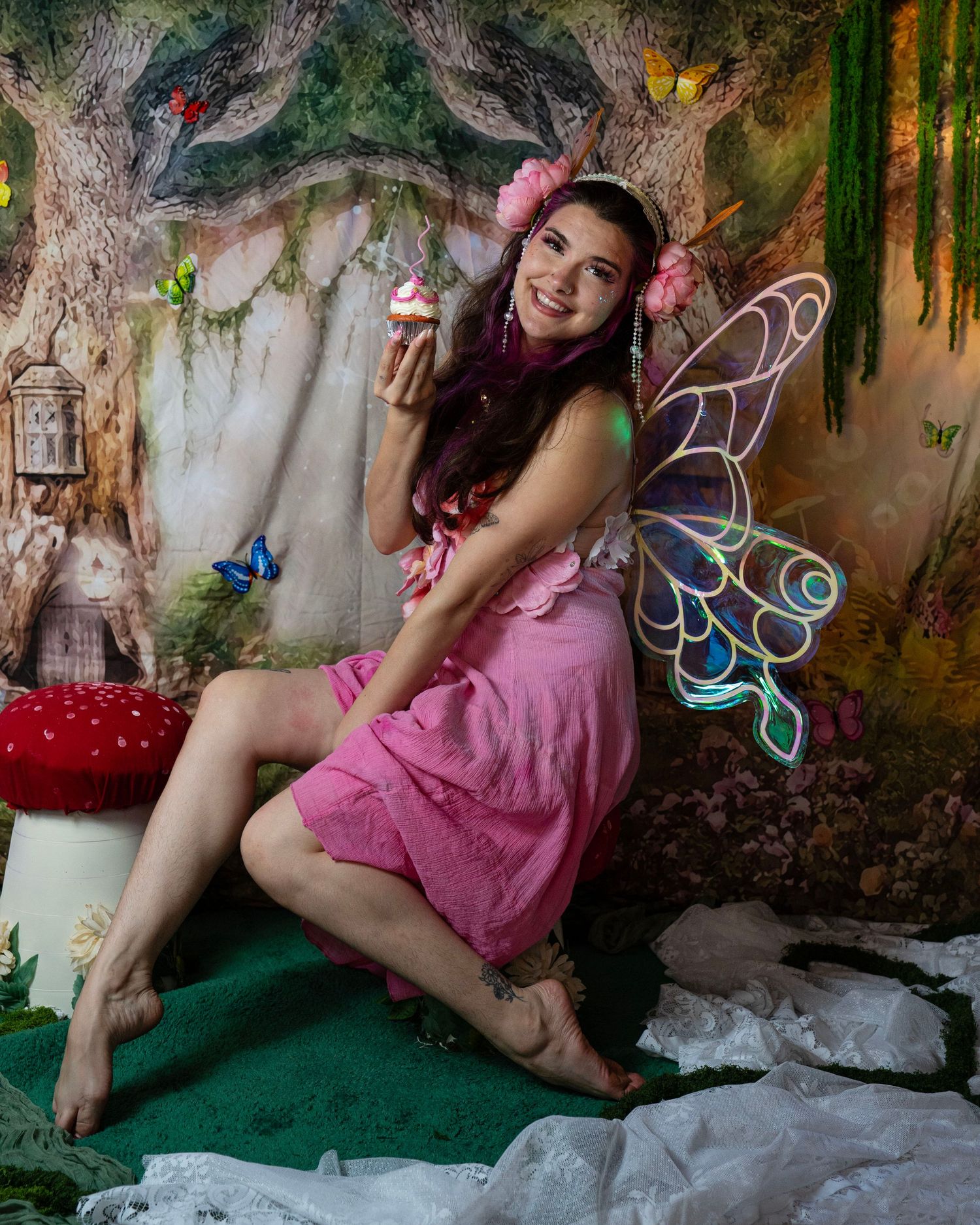 Joyful woman in pink fairy costume with purple iridescent wings holds cupcake beside red mushroom in fantasy setting. Fantasy Photoshoot