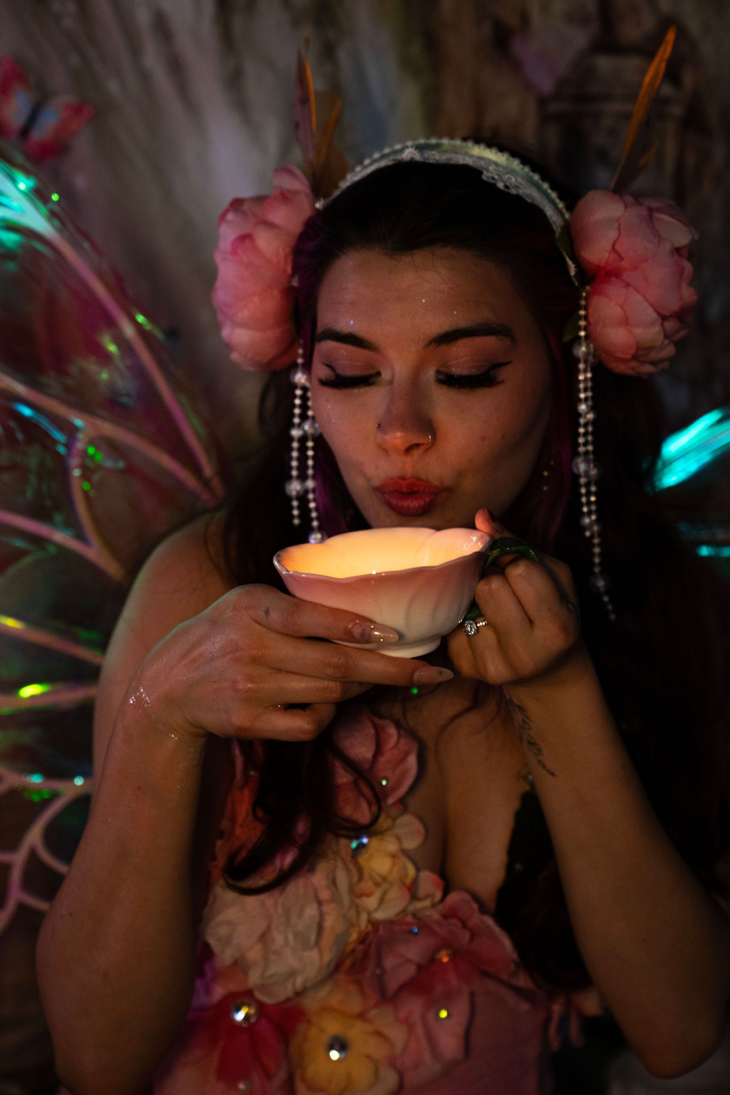 Woman in fairy costume with pink floral headpiece blows on glowing lotus candle in enchanted setting. Fantasy Photoshoot