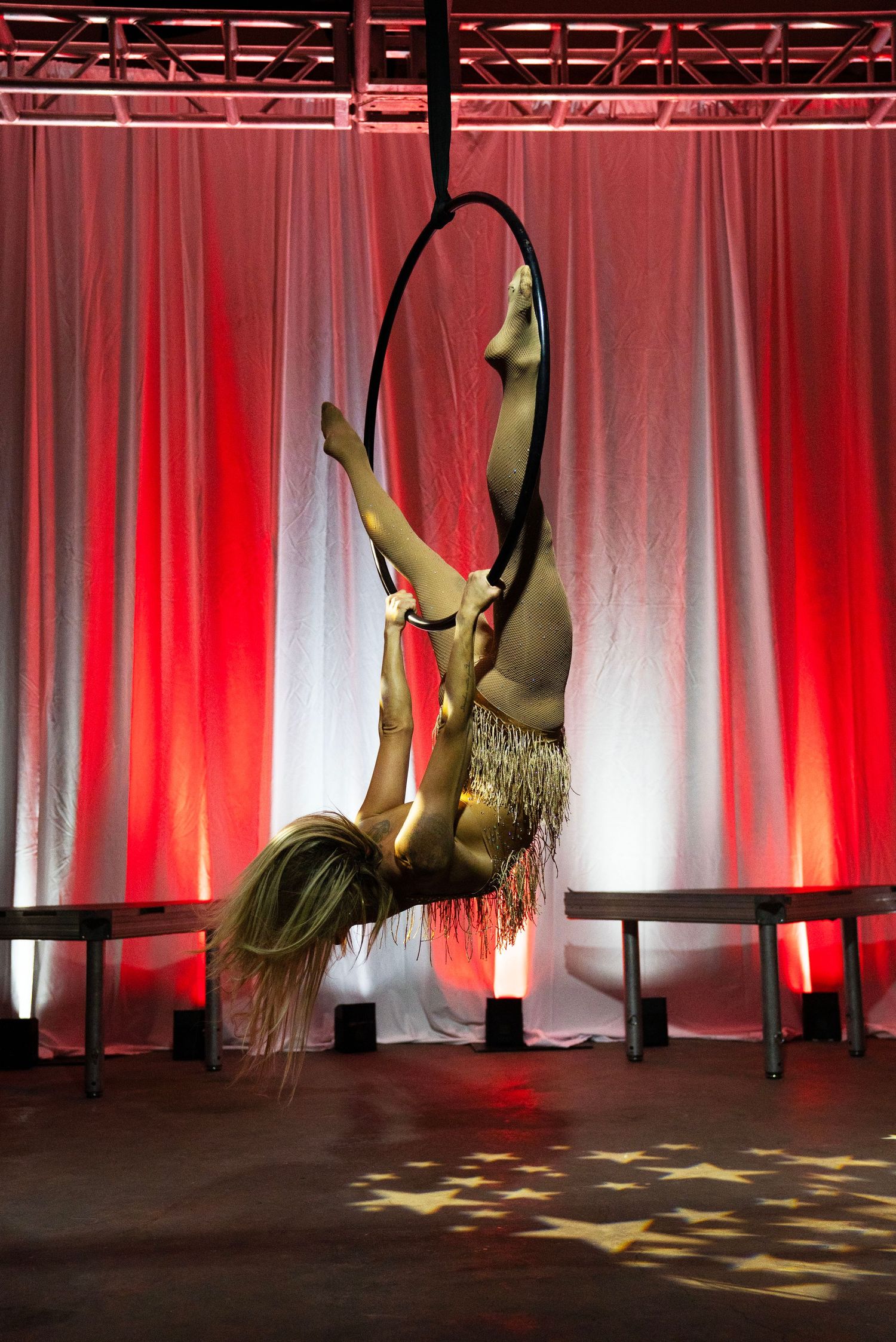 Aerial performer inverted on lyra hoop in gold fringe costume under warm red stage lighting.