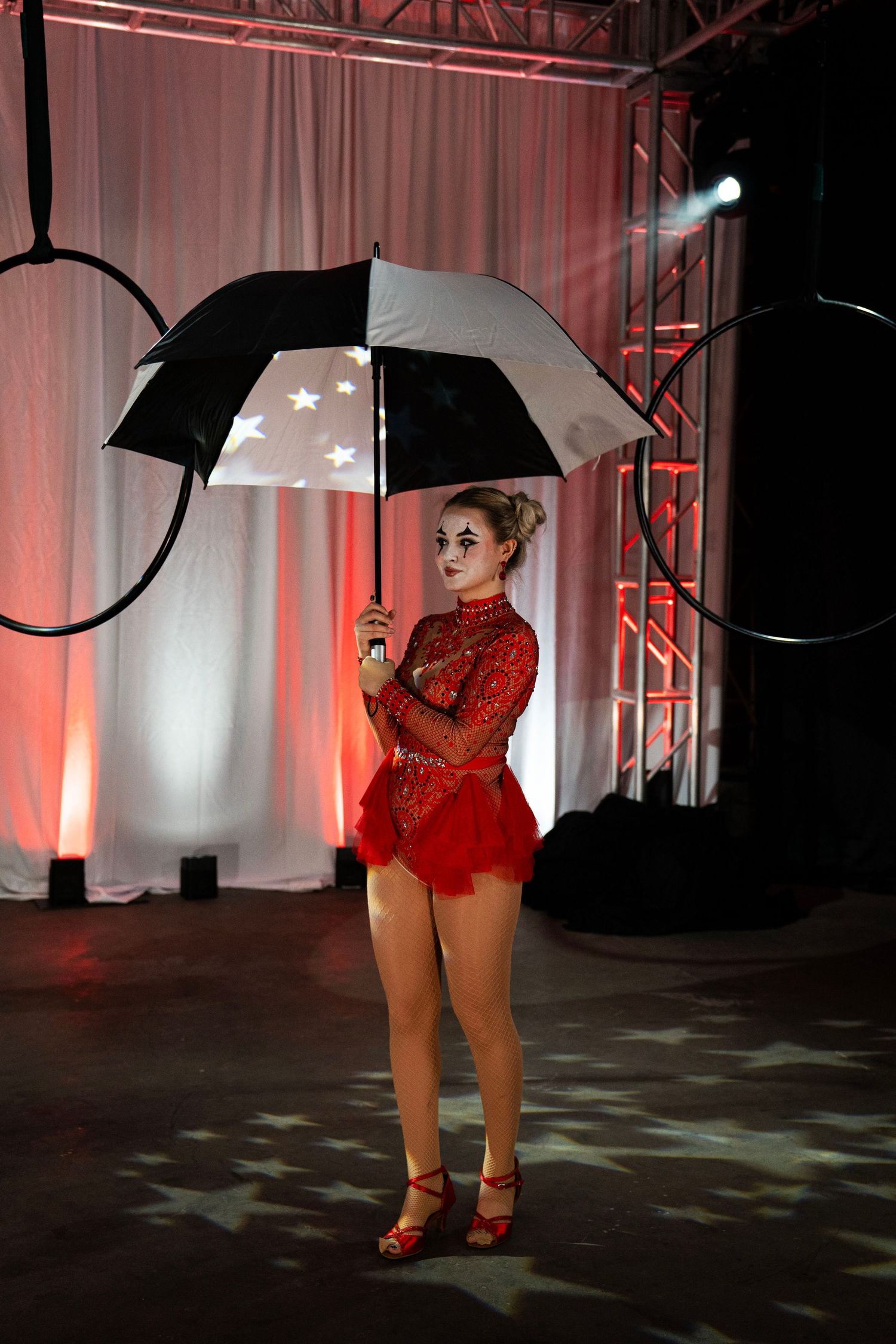 A circus clown in red sequined dress stands holding a black and white star-lined umbrella on stage.