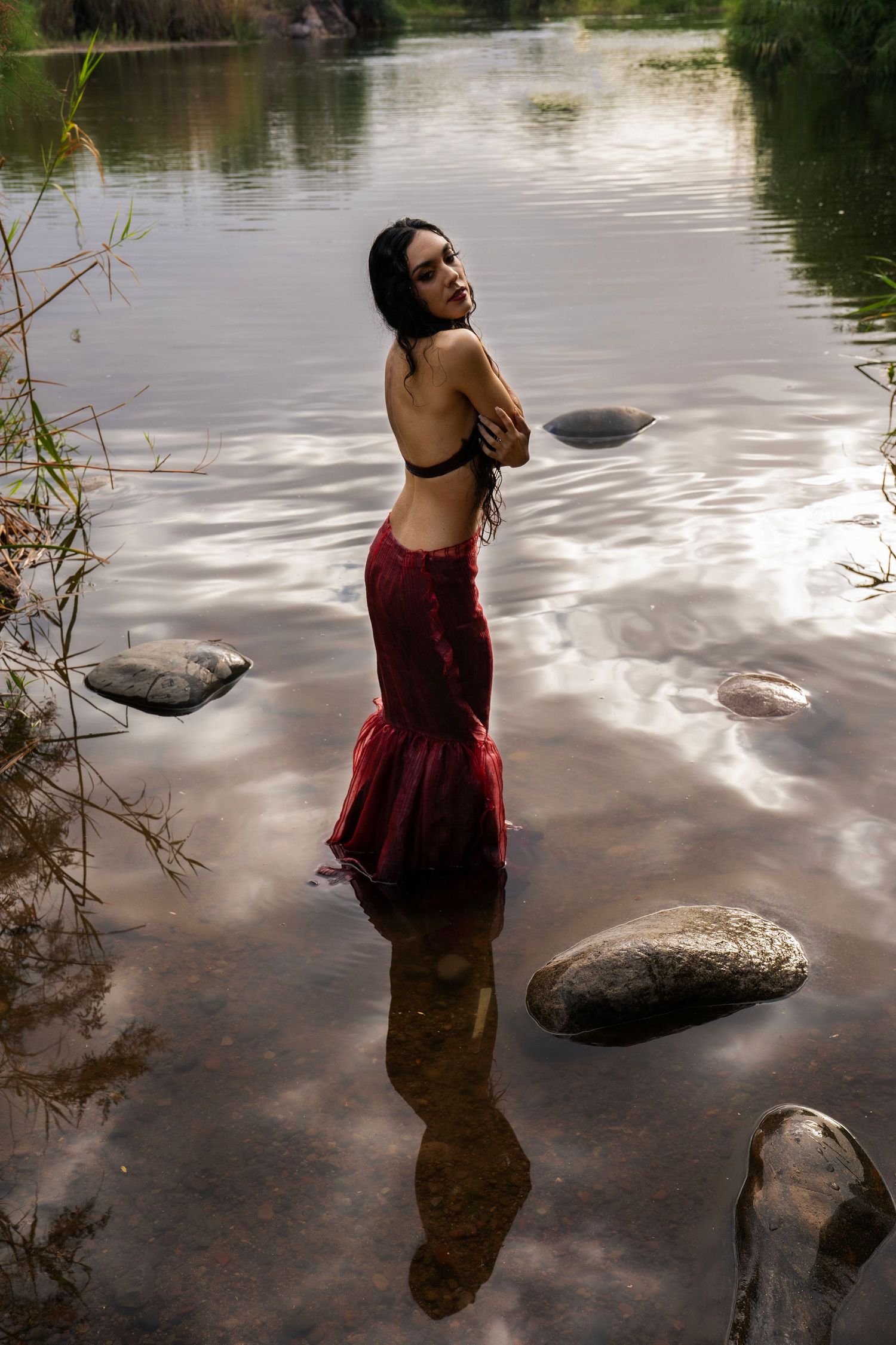 A woman in a red mermaid gown stands in a shallow river, posing with her reflection visible in the calm water.