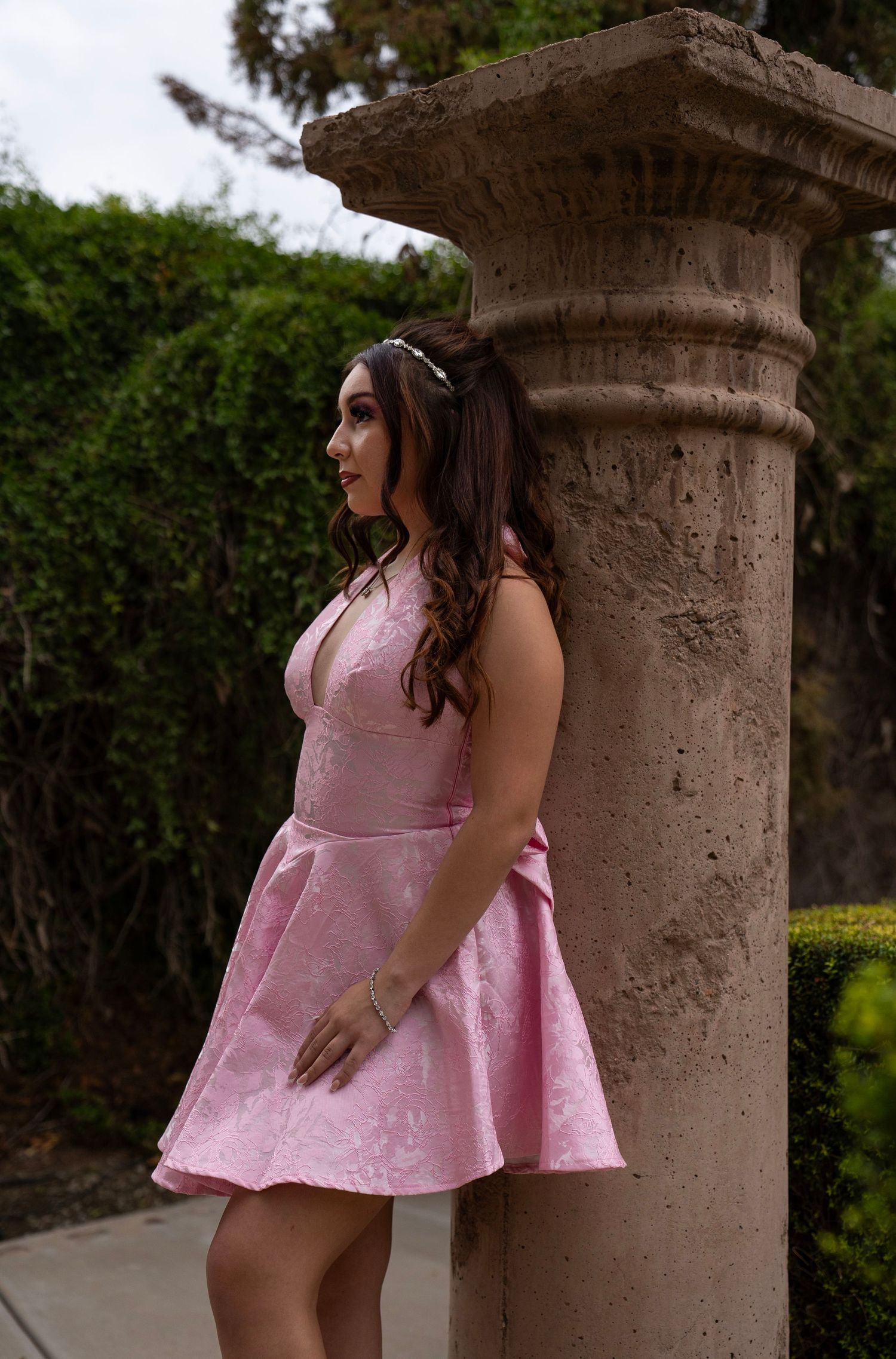 Young woman in pink floral dress leaning against stone column outdoors.