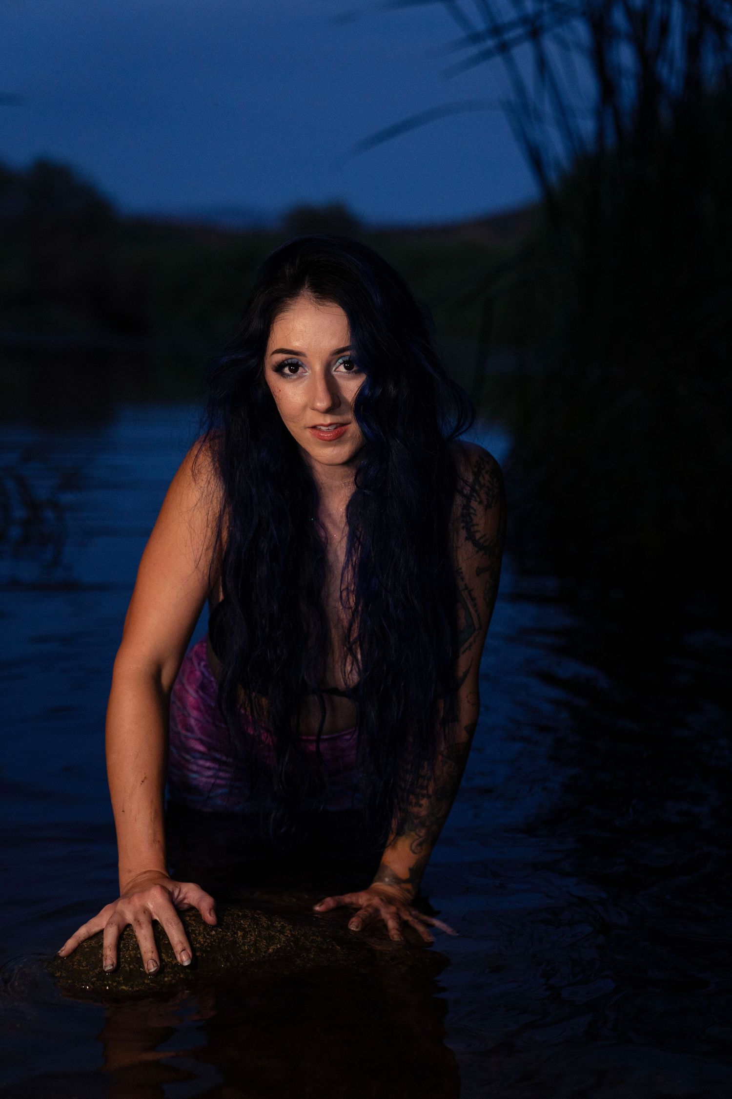 Tattooed woman with long dark hair emerging from a river at dusk in moody blue-toned photography.
