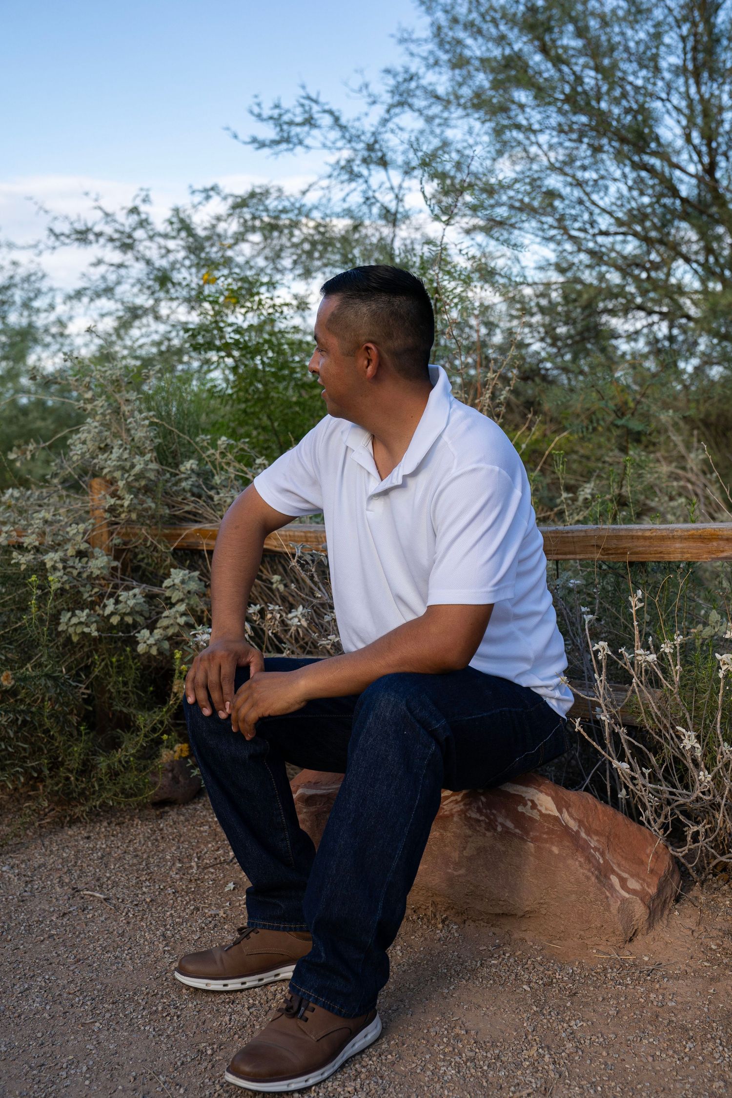Man in white polo shirt and jeans sitting on a rock outdoors, surrounded by desert shrubs and trees.