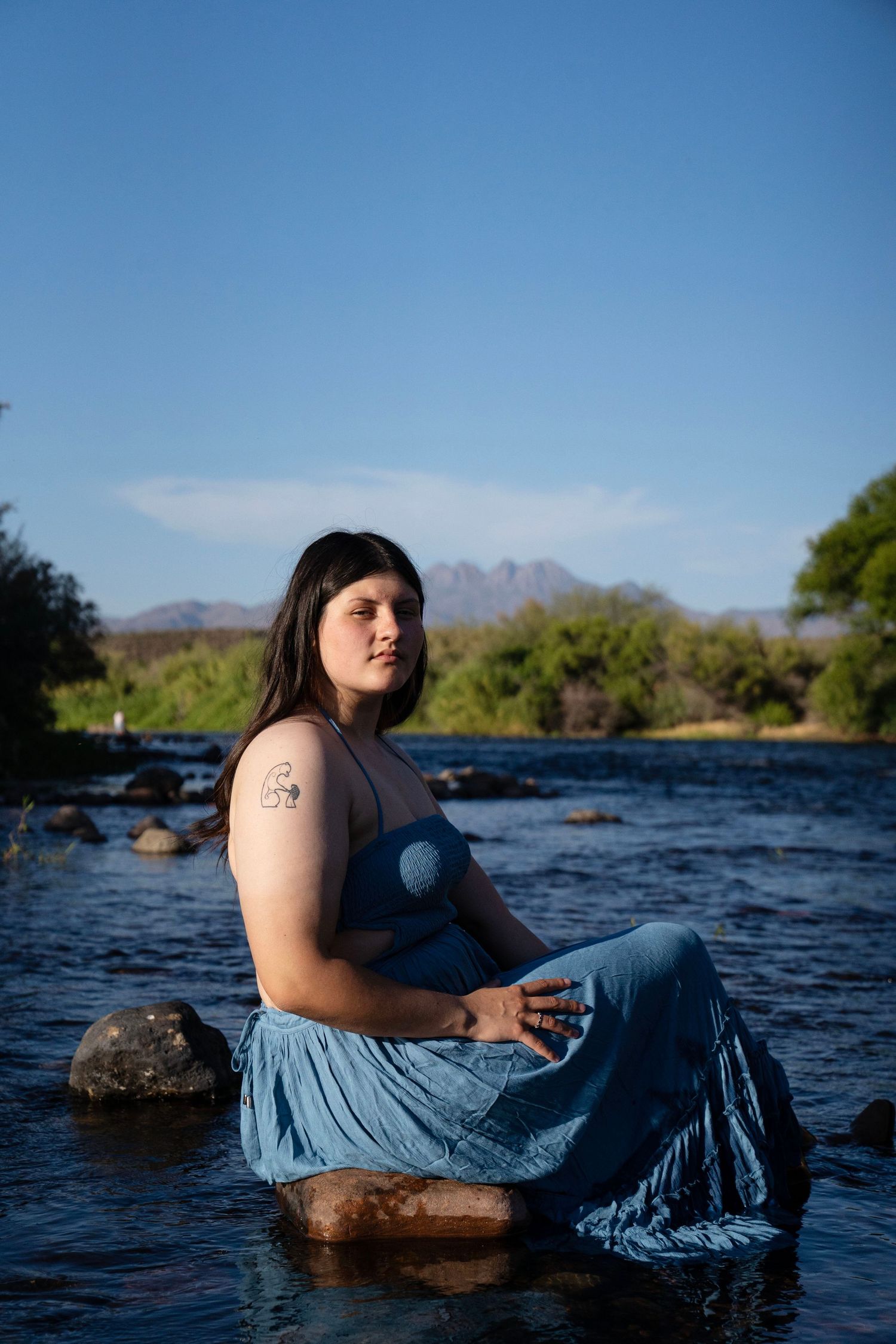 Woman in blue dress sits on river rocks with mountain backdrop under clear blue sky.