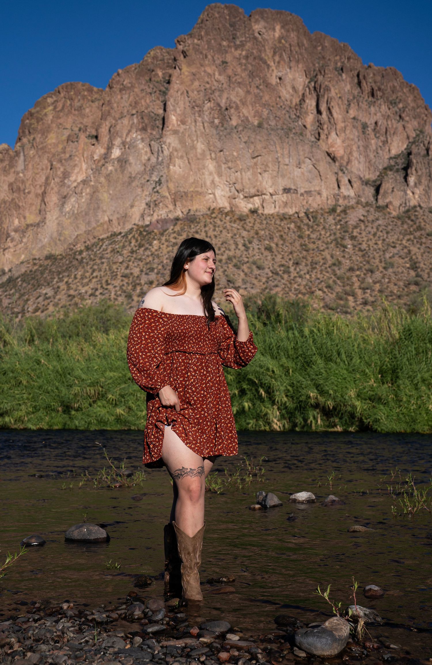 Woman in red floral off-shoulder dress and cowboy boots standing in a rocky river with a mountain backdrop.