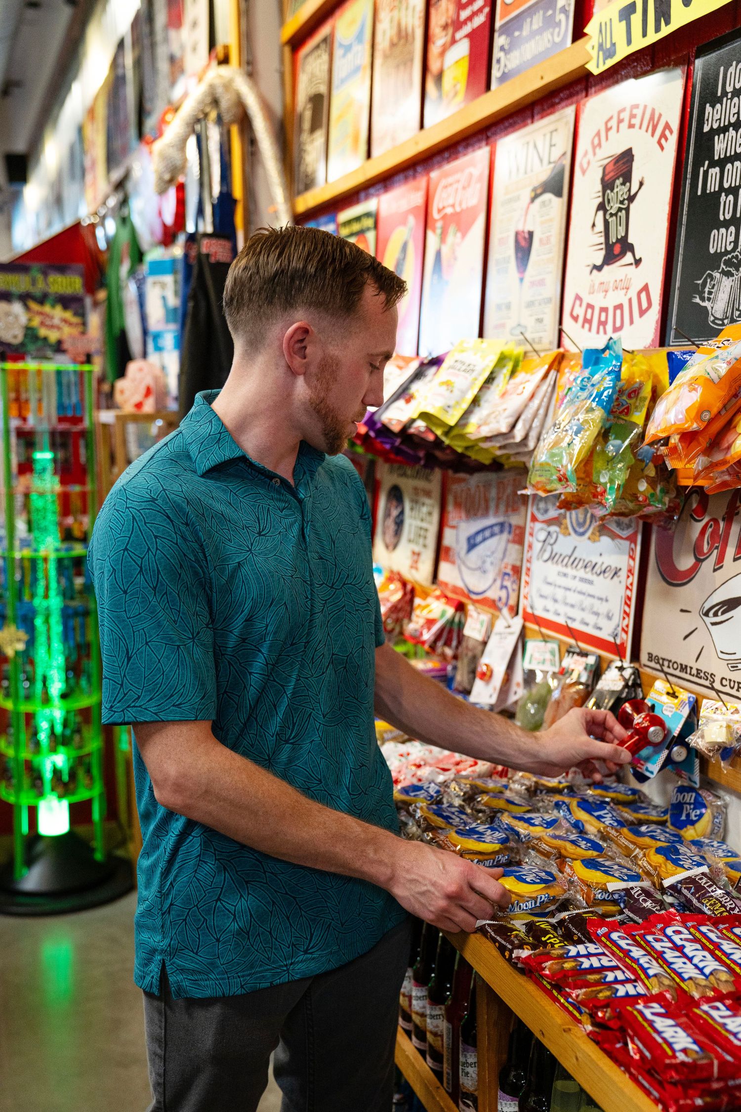 Man in teal shirt browsing candy and novelty items in a colorful retail store with vintage signs.