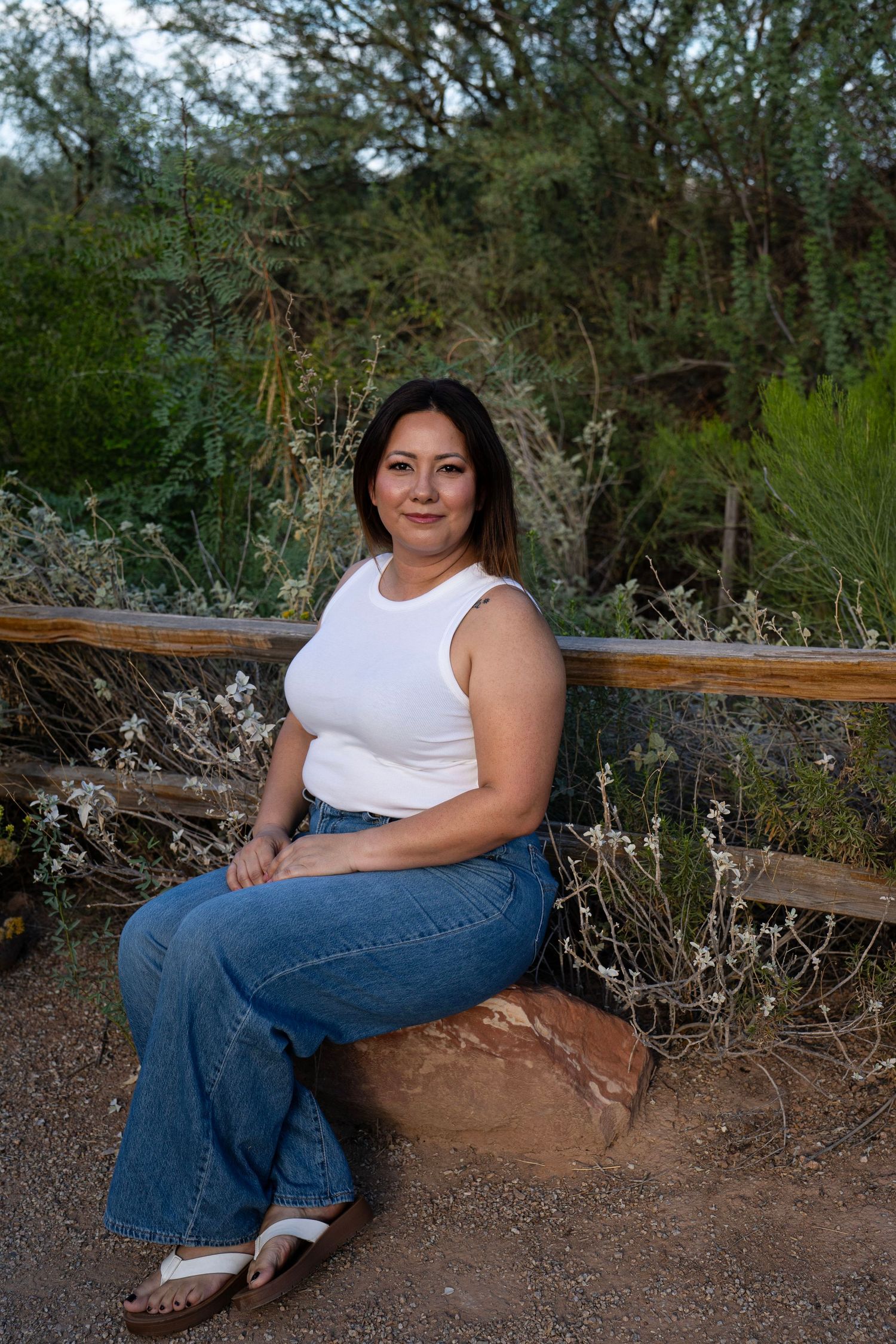 A woman in a white tank top and jeans sits on a rock by a wooden fence surrounded by lush desert greenery.