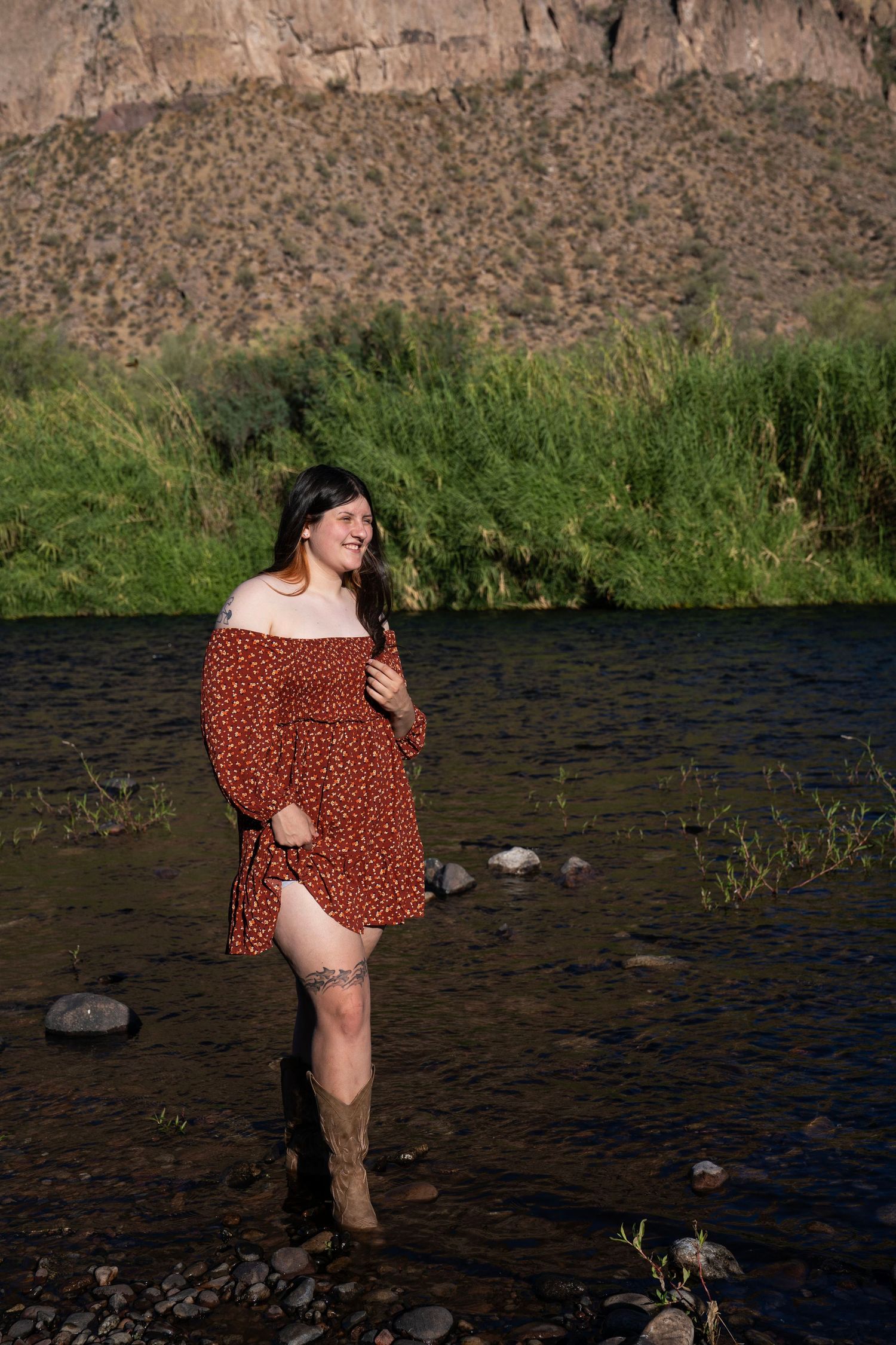 Woman in red floral dress and cowboy boots standing in a rocky river with green hills in background.