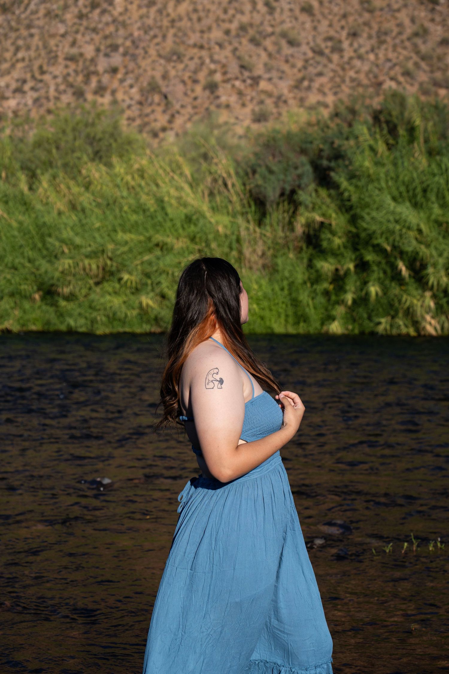 Woman in blue maxi dress standing by a river with green vegetation and rocky hills in the background.
