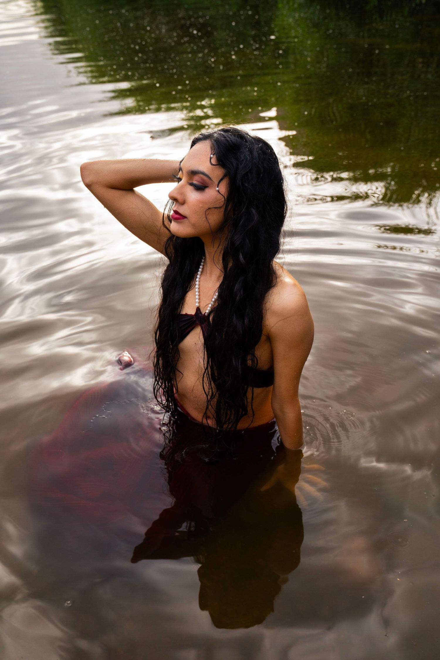 A woman with long wet dark hair and bold makeup poses in a river wearing a pearl necklace and dark top.