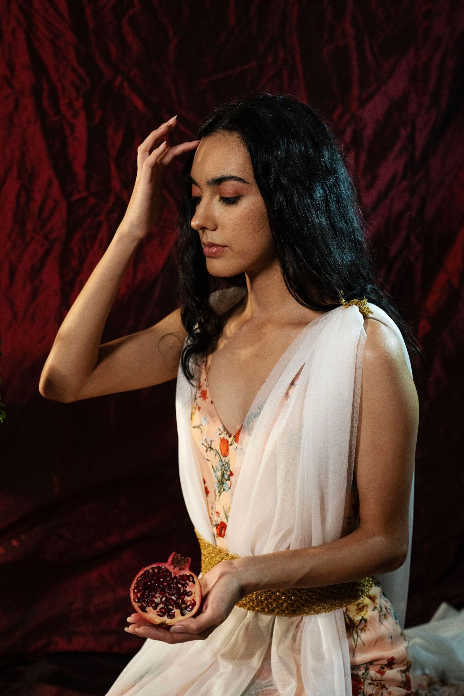 Woman in a white draped gown with a gold belt holds a halved pomegranate and touches her forehead against a deep red velvet backdrop.