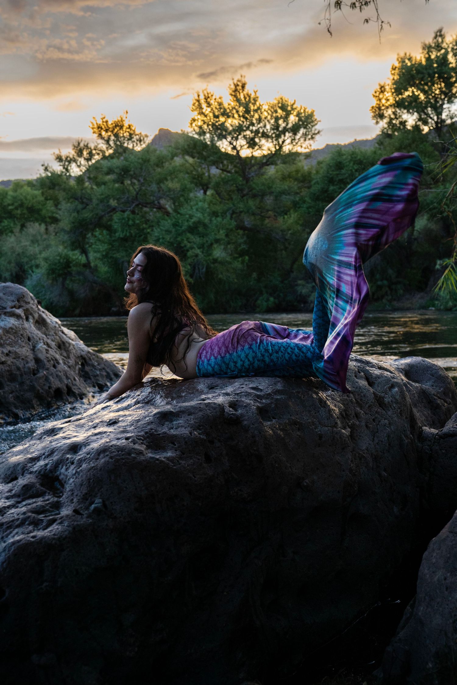 Woman in colorful mermaid tail costume reclines on rock by river at golden sunset. Mermaid photoshoot, fantasy photoshoot.