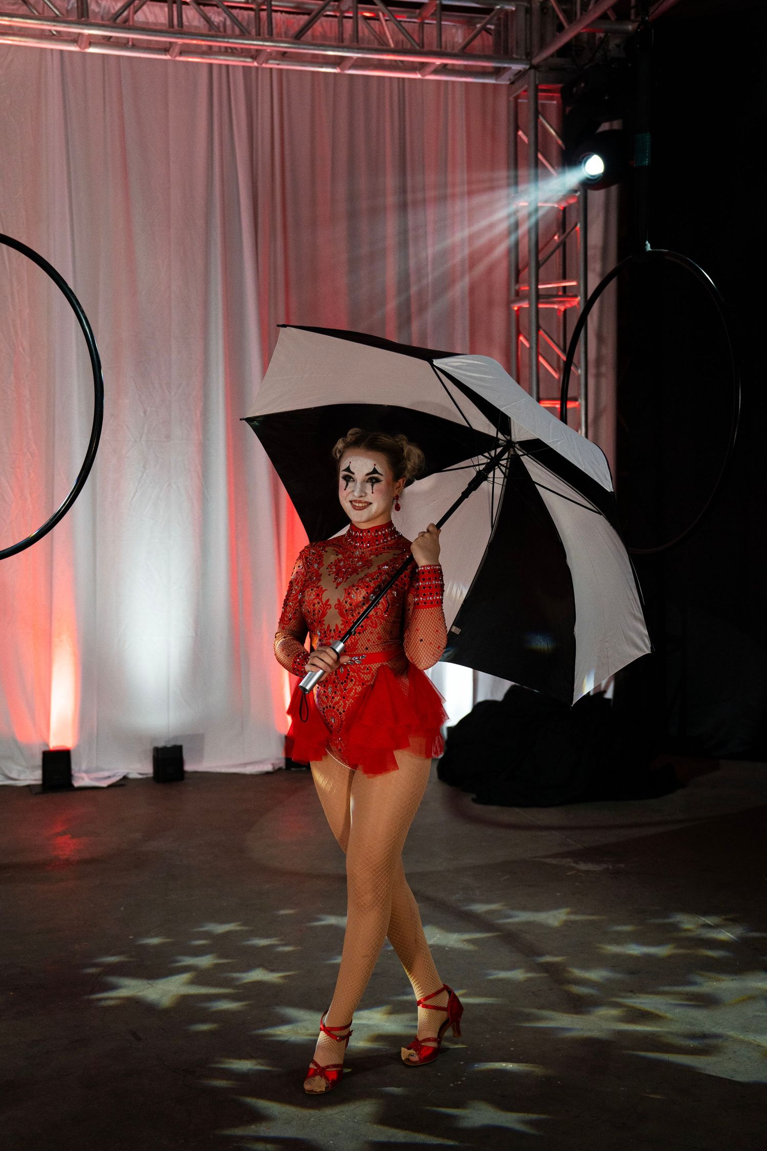 A circus performer in red costume with clown makeup walks holding a black and white umbrella on stage.