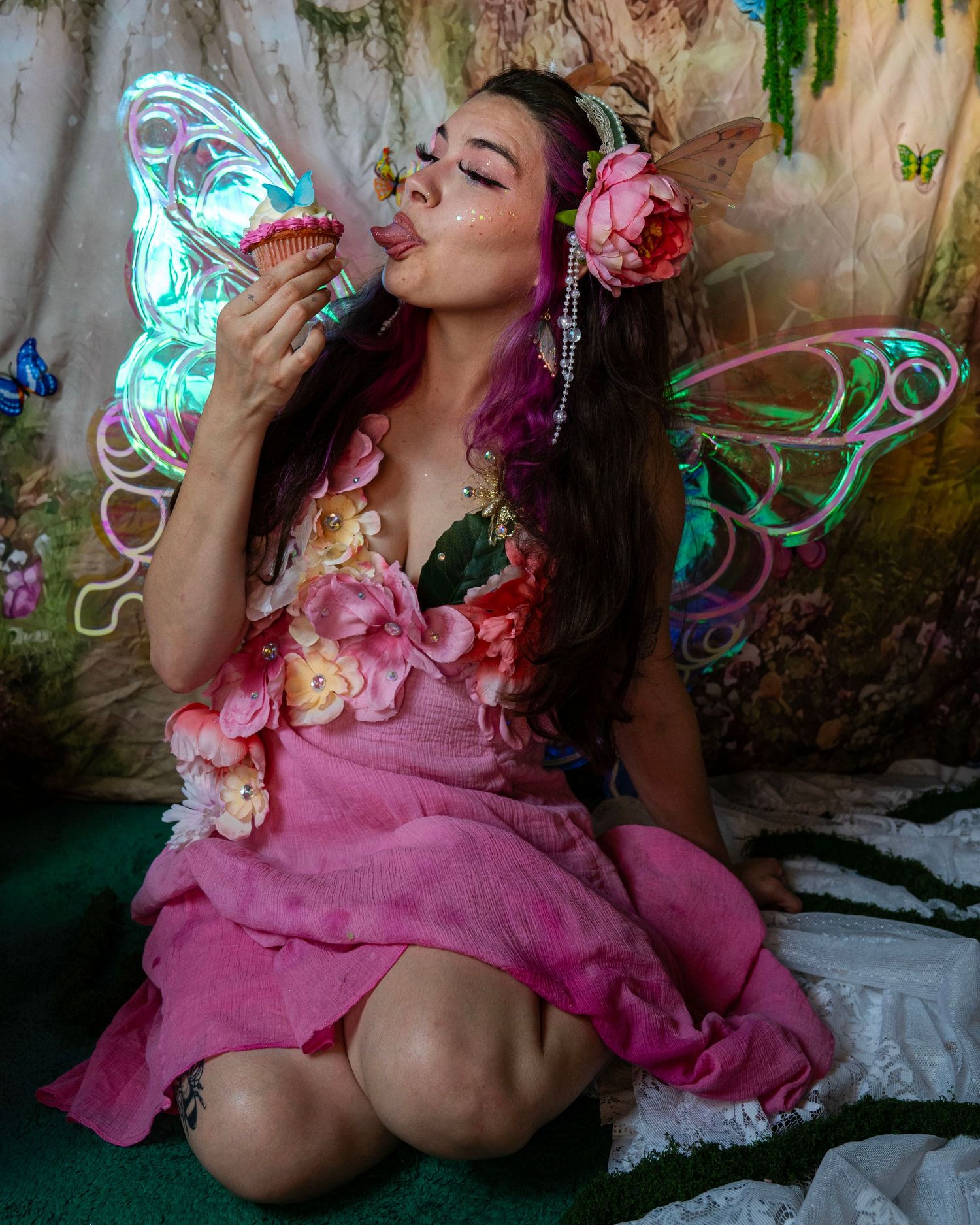 Woman in pink fairy dress with illuminated wings licks frosting from a cupcake in an enchanted forest setting. Fantasy Photoshoot