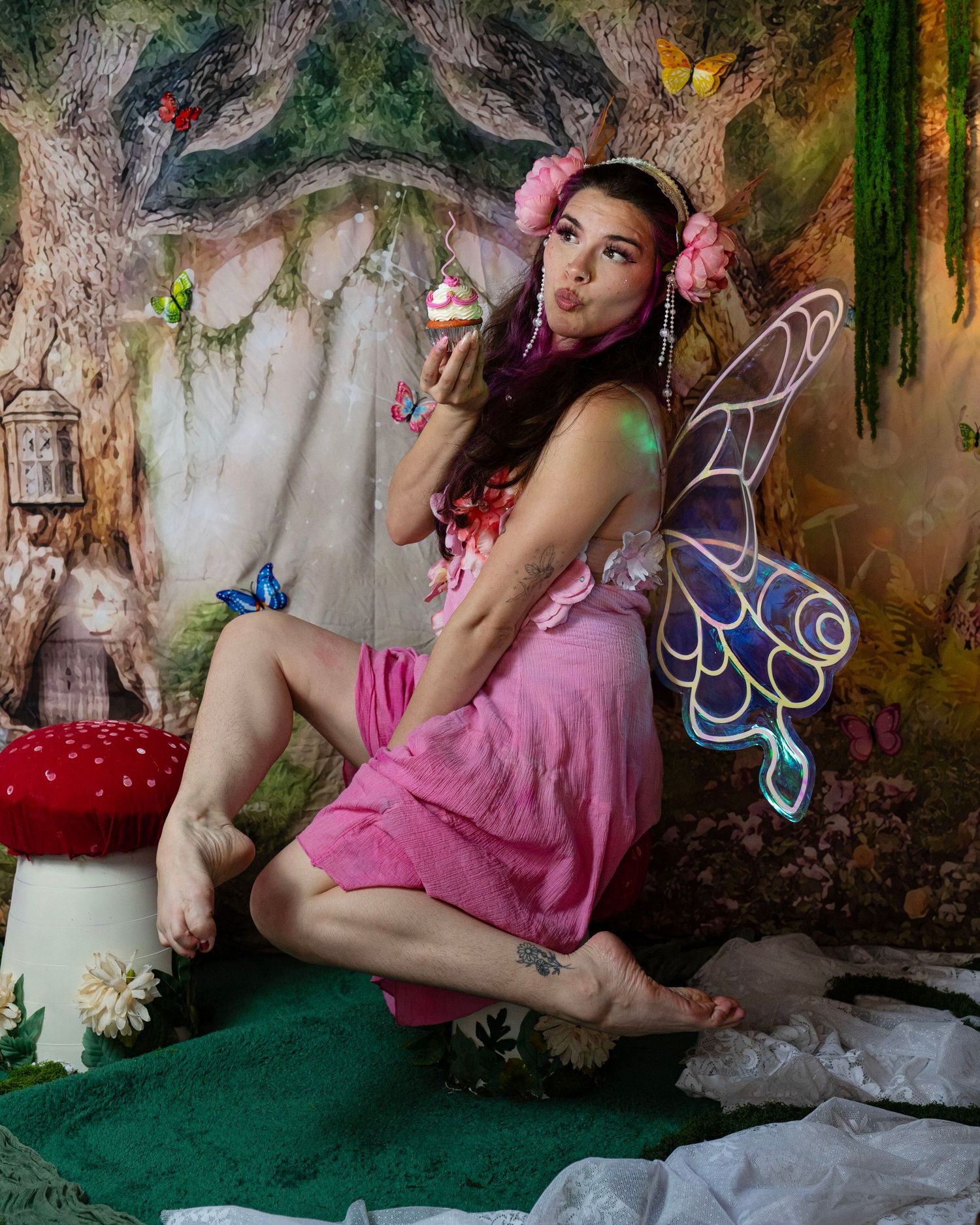 Woman in pink fairy dress with decorated wings blows kiss while holding cupcake in whimsical forest photo studio. Fantasy Photoshoot