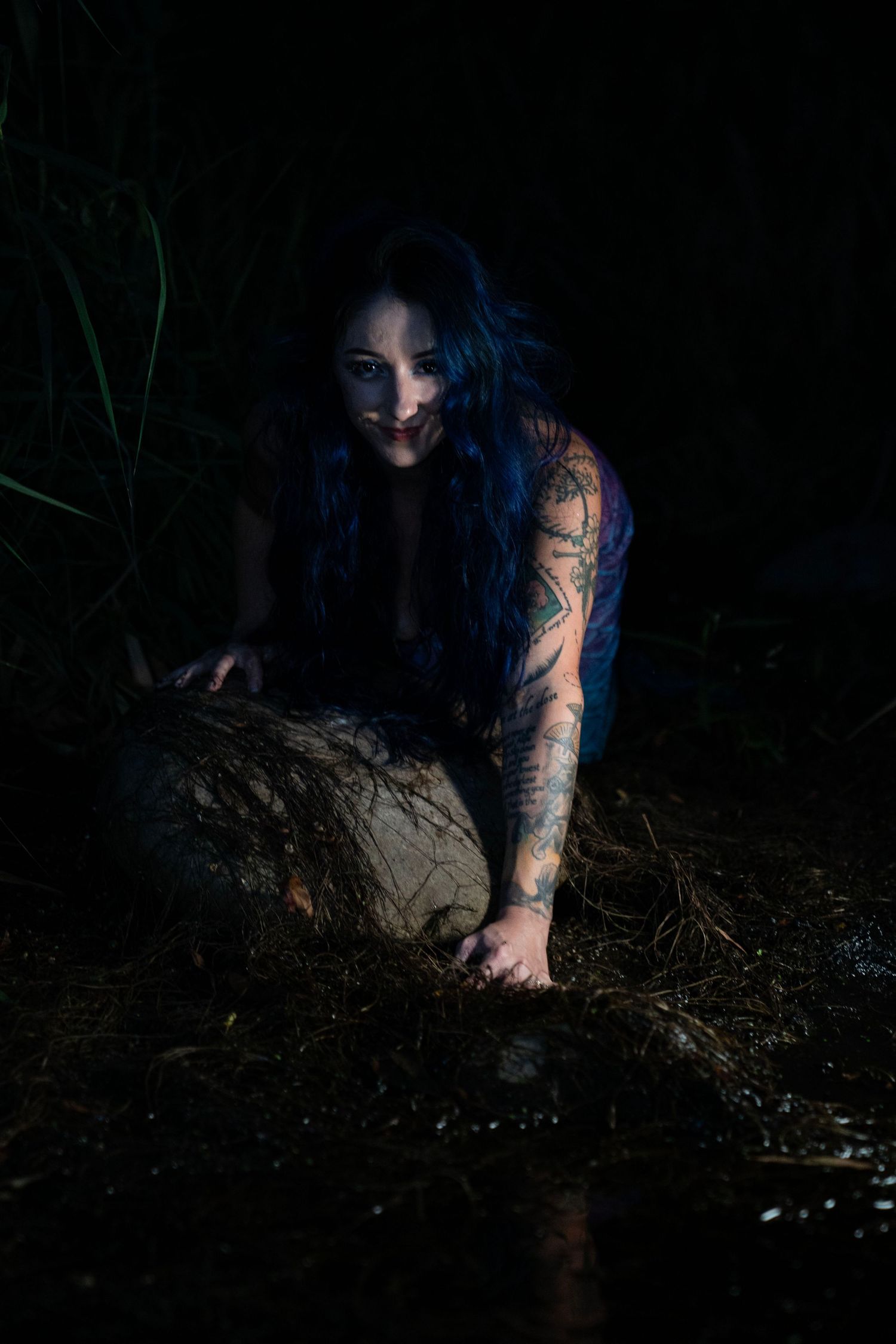 Tattooed woman with blue hair crouches by rock in dark outdoor setting.