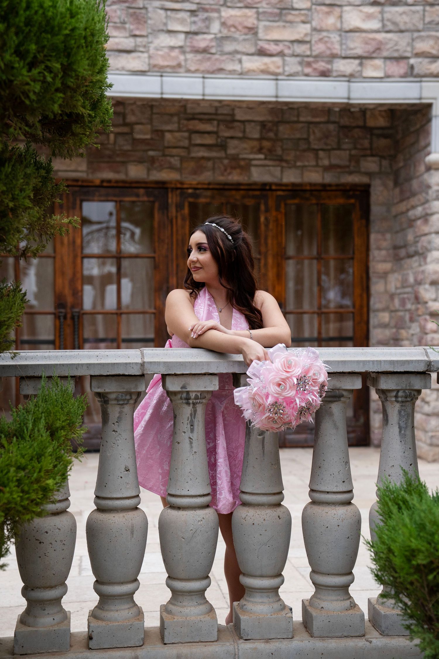 Woman in pink dress leaning on stone balustrade holding a pink bouquet outside an elegant stone building.