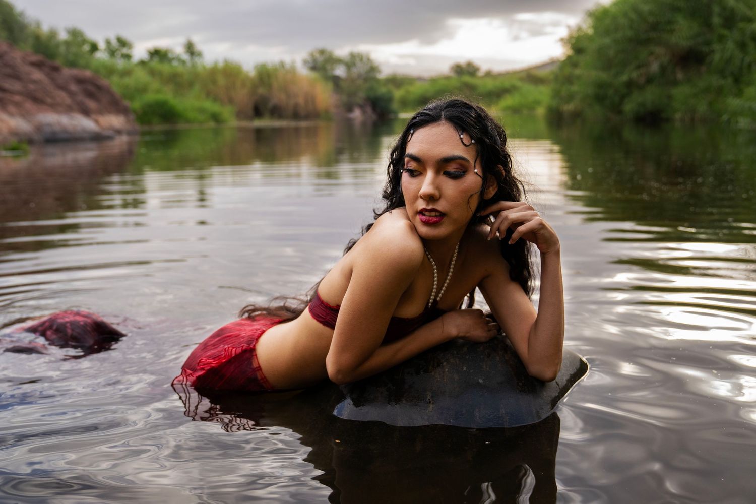 A woman in a red outfit poses on a rock in a river, leaning forward with wet hair in a mermaid-inspired photoshoot.