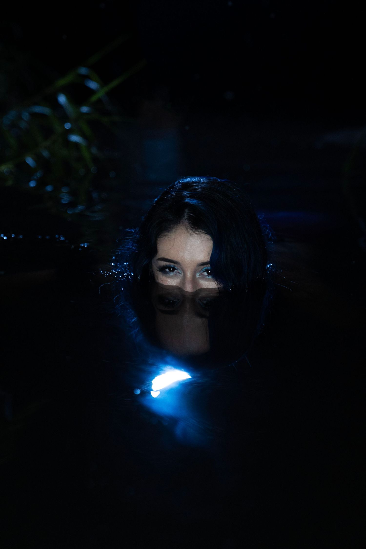 A mysterious woman submerged in dark water at night, her eyes visible above the surface with a blue light glowing nearby.