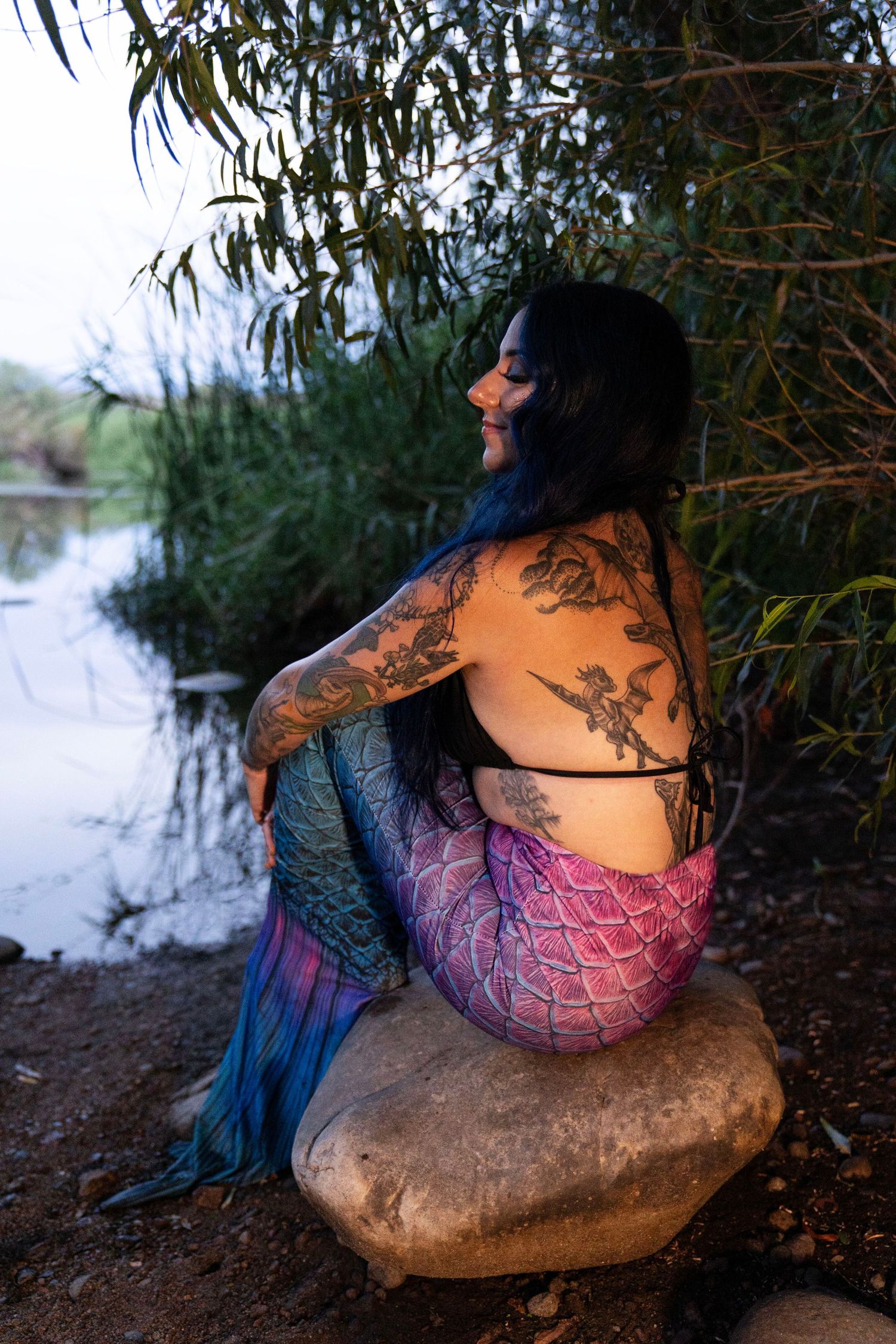 Tattooed woman in colorful mermaid tail costume sitting on rocks by a river at dusk.