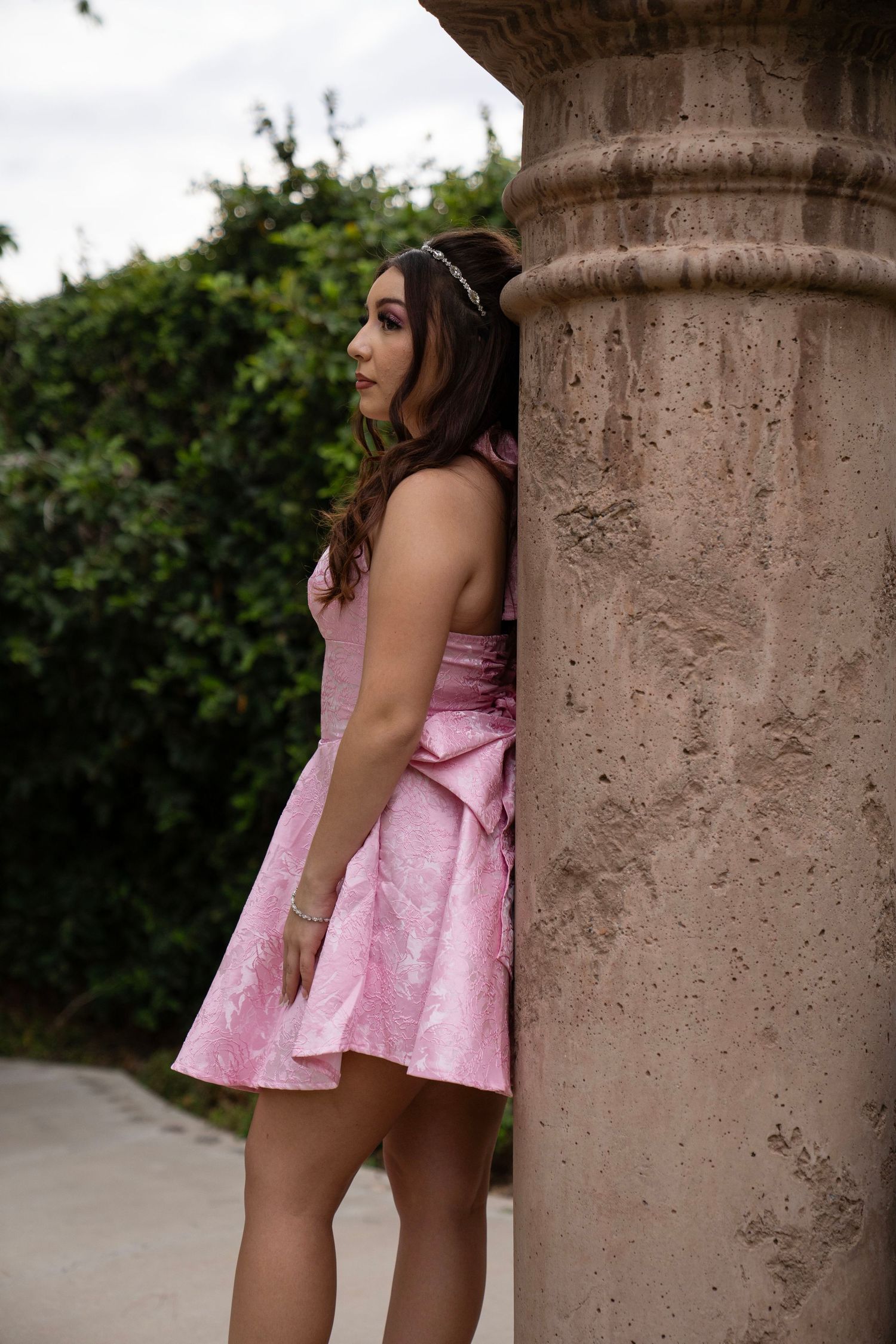 Young woman in pink formal dress with bow poses beside stone column outdoors.