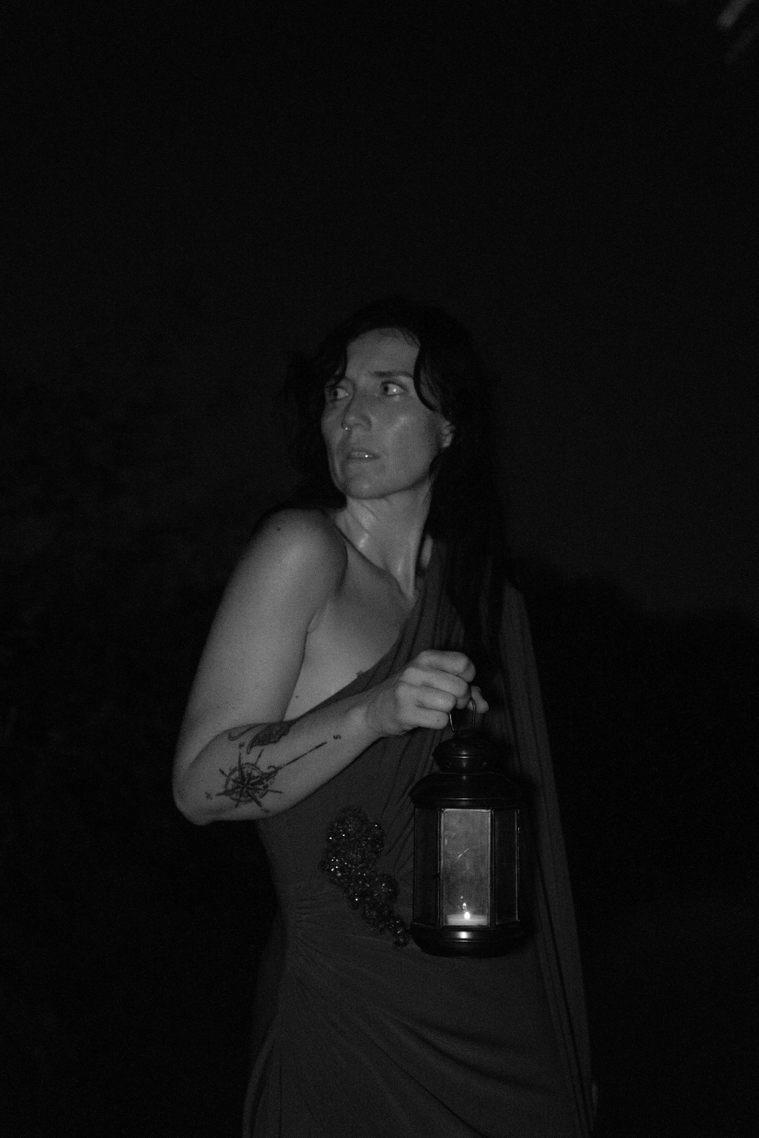 Black and white portrait of a woman with a floral arm tattoo holding a lit lantern in the dark, glancing over her shoulder.