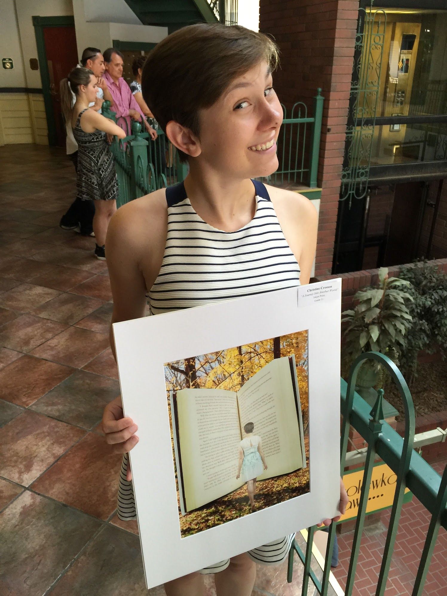 Smiling woman in a striped sleeveless top holds a framed print of a girl standing before a giant open book in autumn woods.