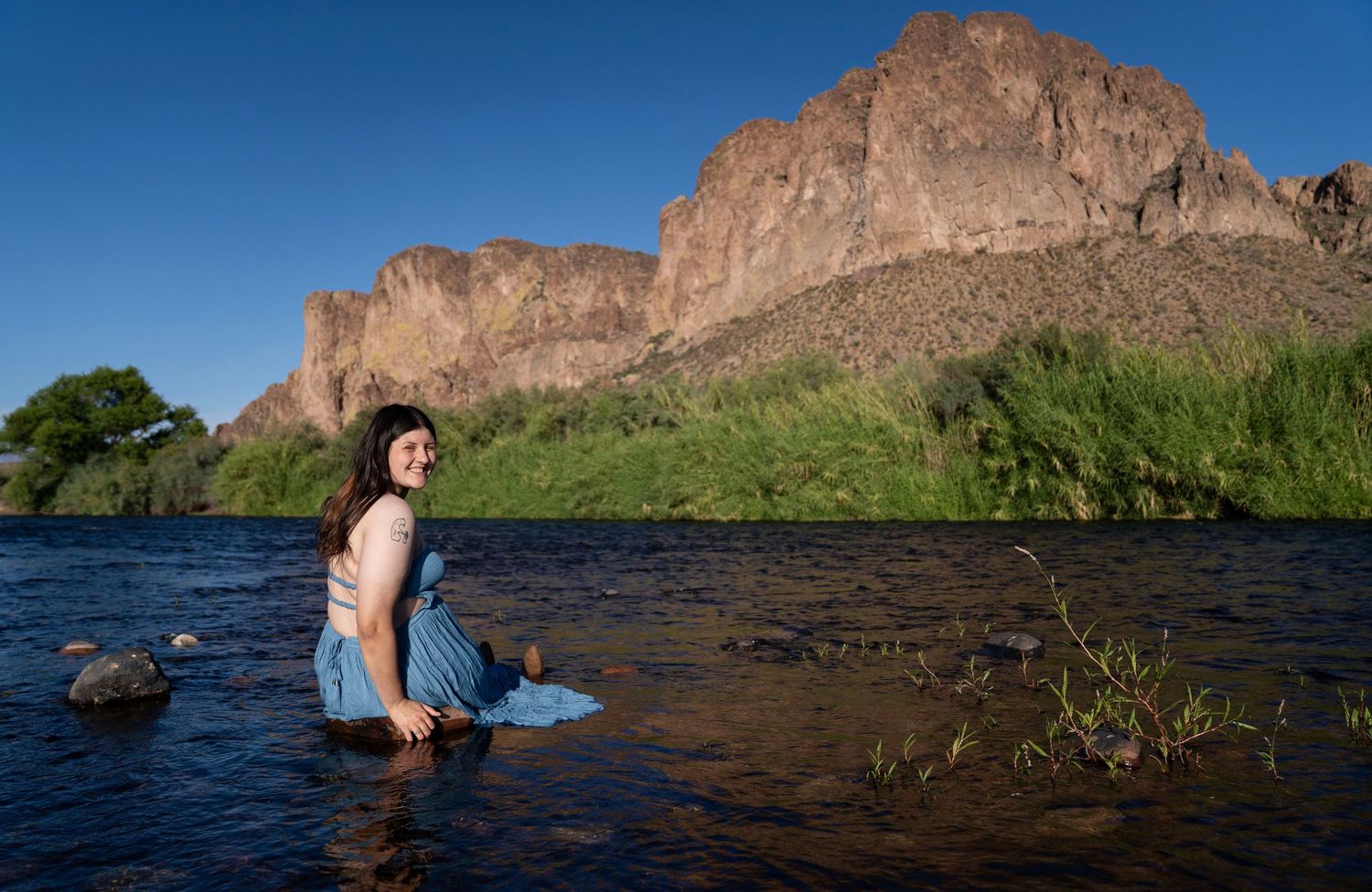 Woman wading in a desert river with dramatic rocky cliffs and green trees under a clear blue sky.