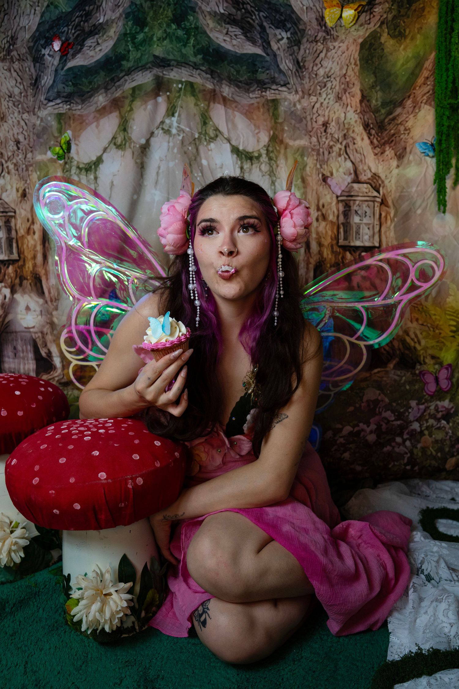 Smiling fairy with iridescent wings and pink floral dress holds a cupcake while seated on a mushroom in an enchanted forest. Fantasy