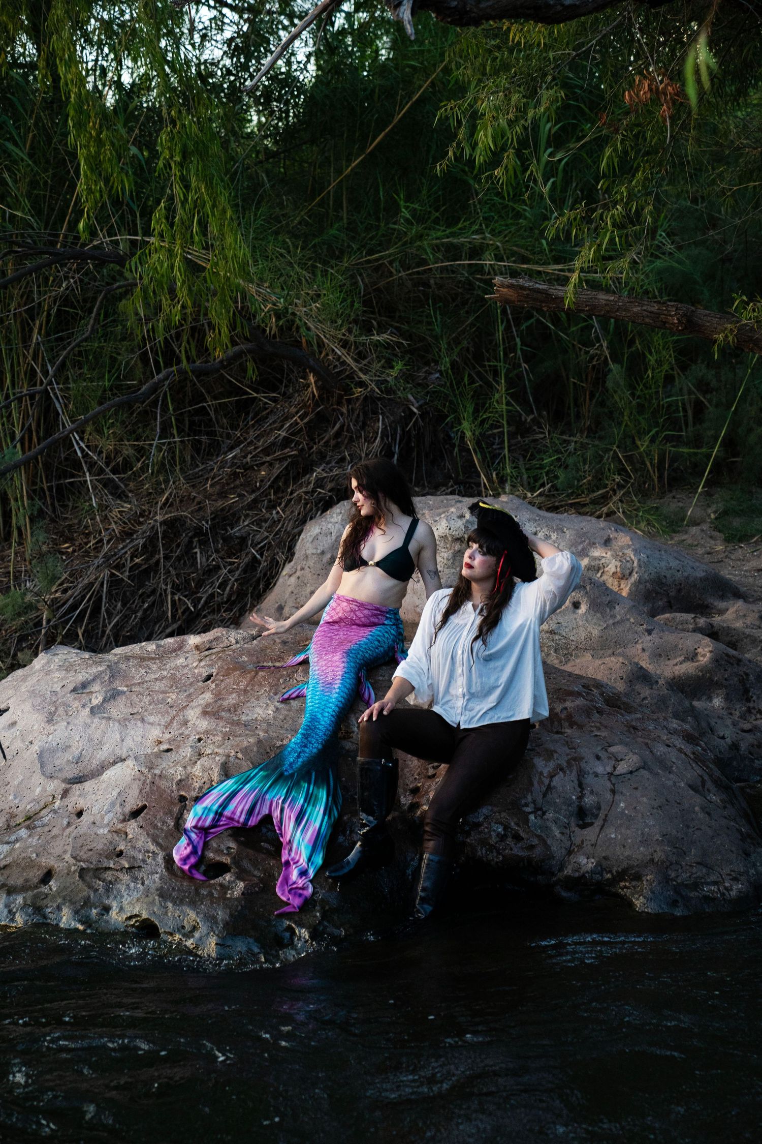 A mermaid and pirate costumed duo pose dramatically on rocks beside a dark, tree-lined river. Mermaid photoshoot, fantasy photoshoot.