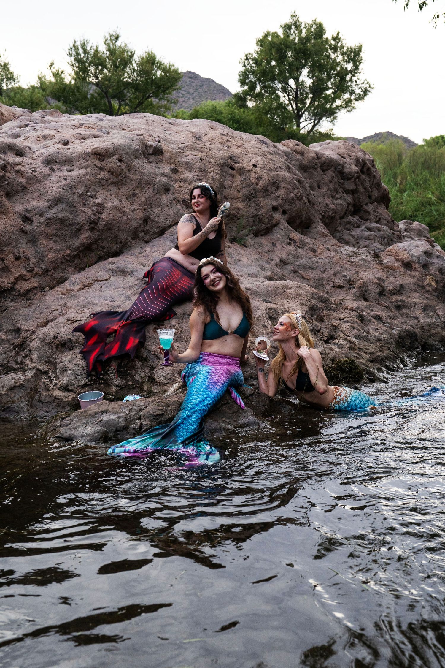 Three women in colorful mermaid costumes pose together on rocks beside a river. Fantasy Photoshoot
