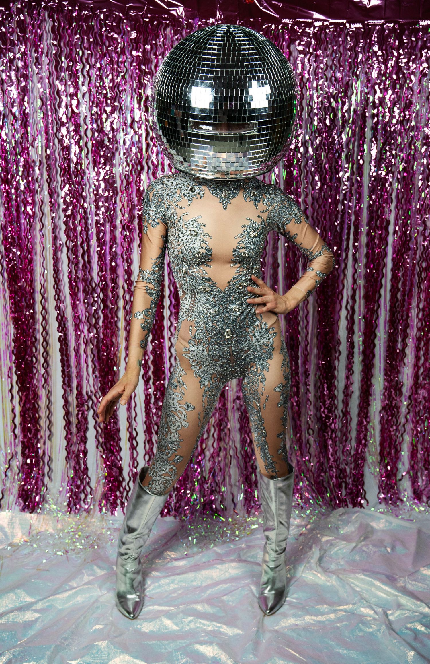 Model wearing disco ball helmet and silver rhinestone catsuit with silver boots on pink backdrop.