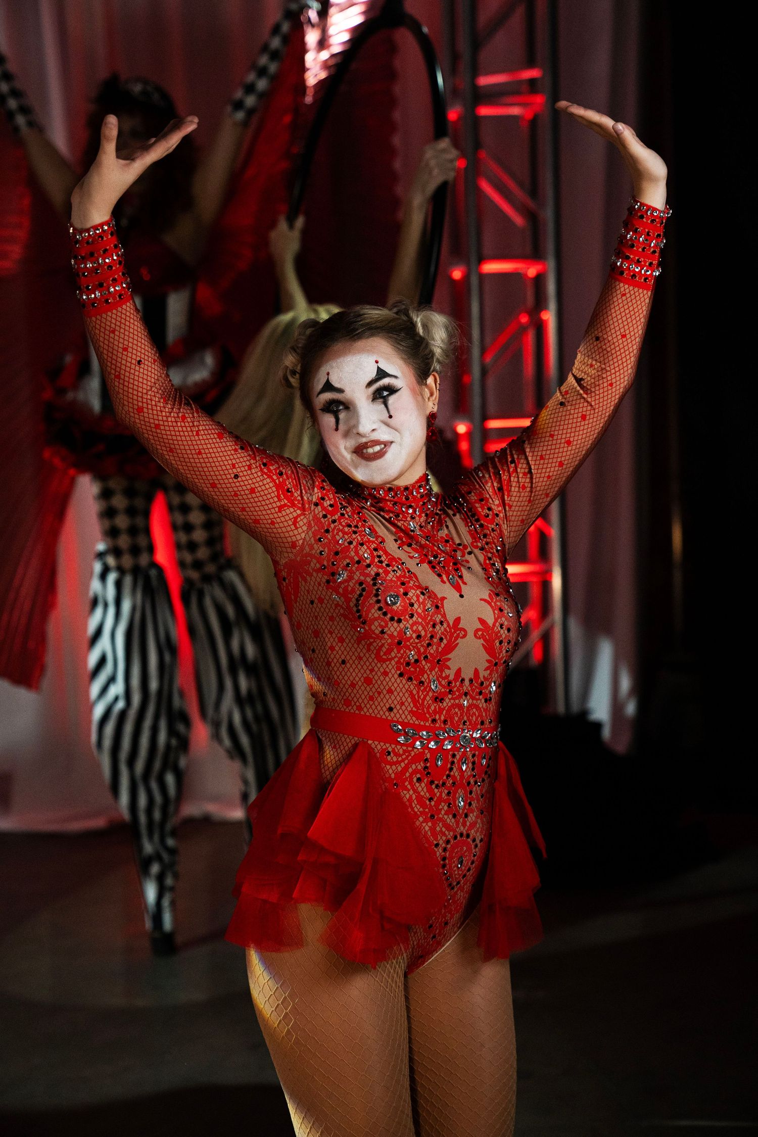 A smiling circus performer in a red jeweled leotard with clown makeup raises her arms on stage.