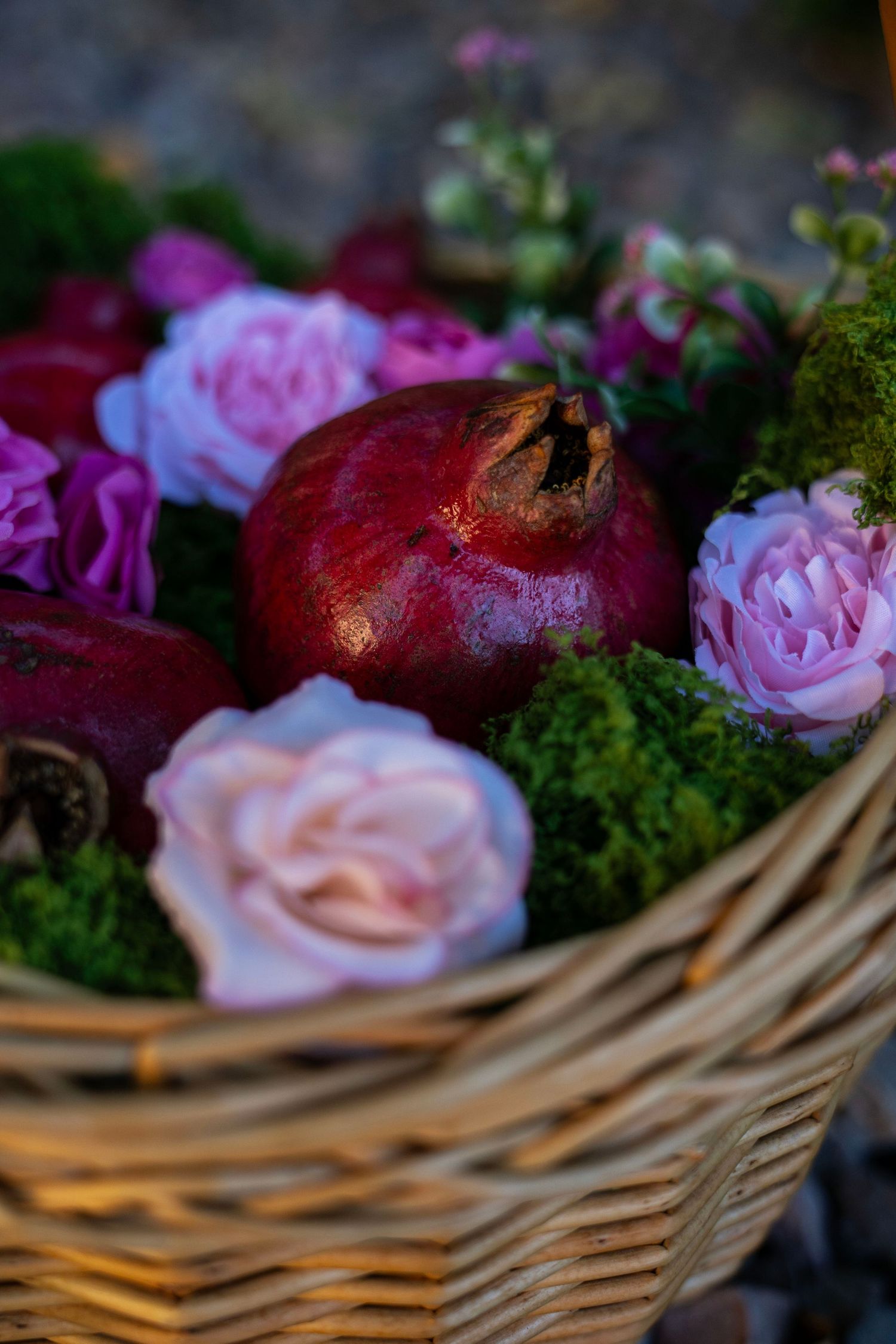 Wicker basket filled with pink roses, deep red pomegranates and green moss, arranged as a rustic autumn floral centerpiece.