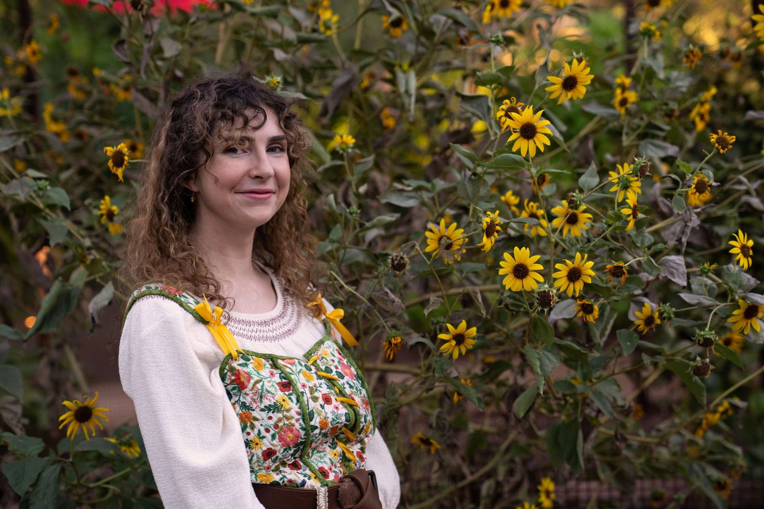Smiling woman with curly hair wears a floral embroidered blouse and beaded necklace, standing in front of blooming yellow sunflowers.
