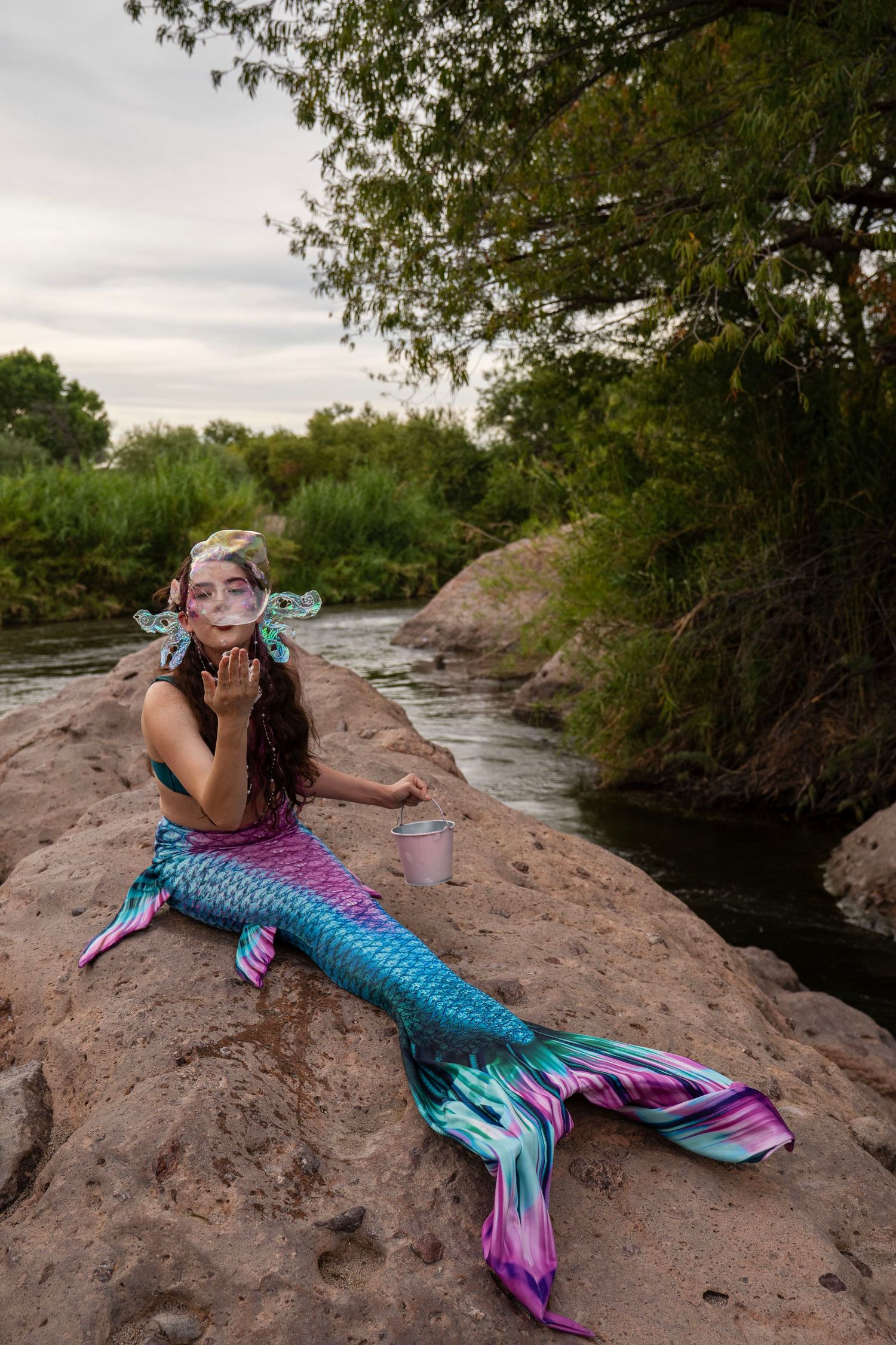 A woman dressed as a mermaid blows bubbles while sitting on rocks near a flowing river. Fantasy Photoshoot