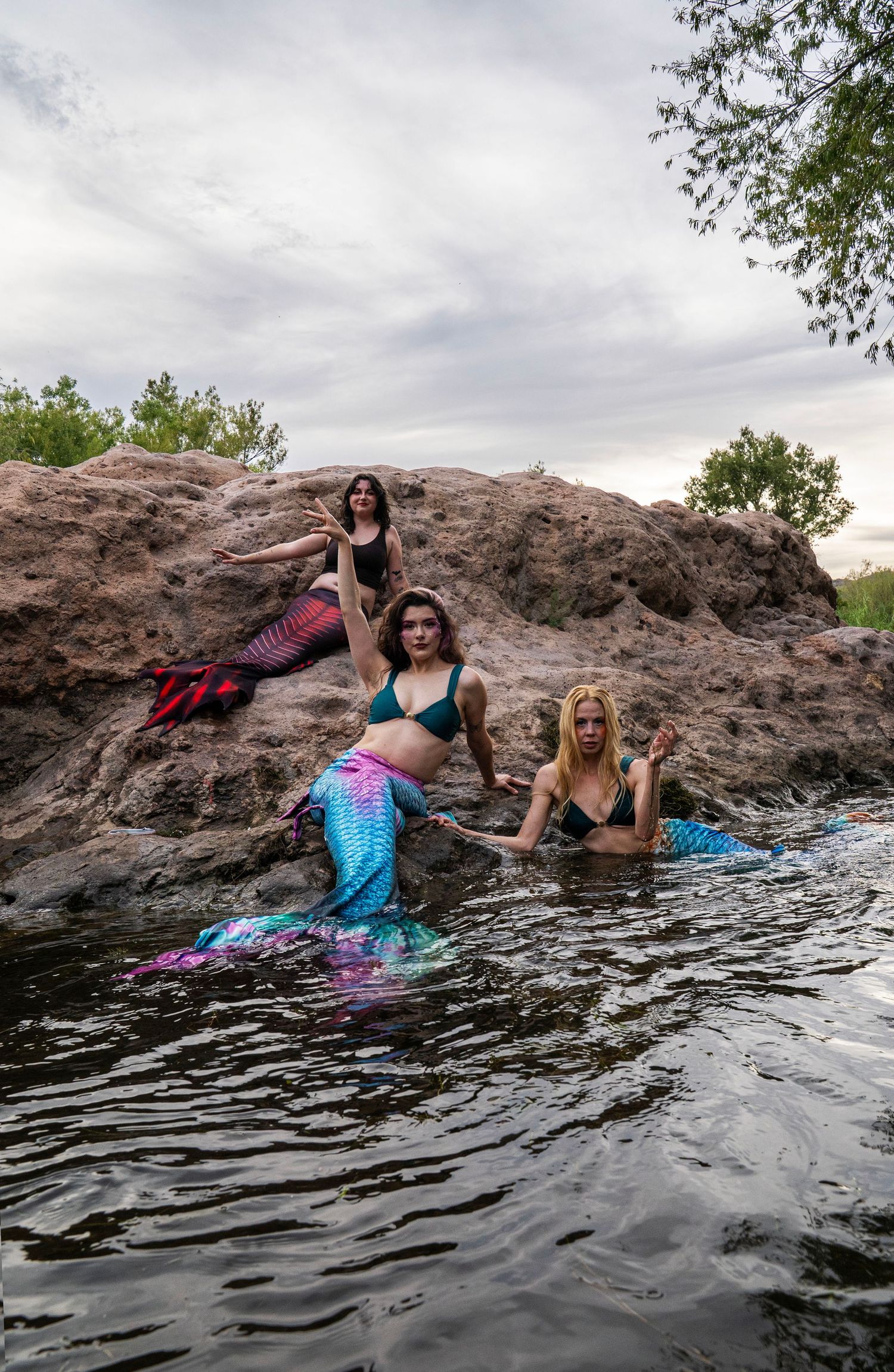 Three women in mermaid costumes with colorful tails pose on rocks and in a river at twilight. Fantasy Photoshoot