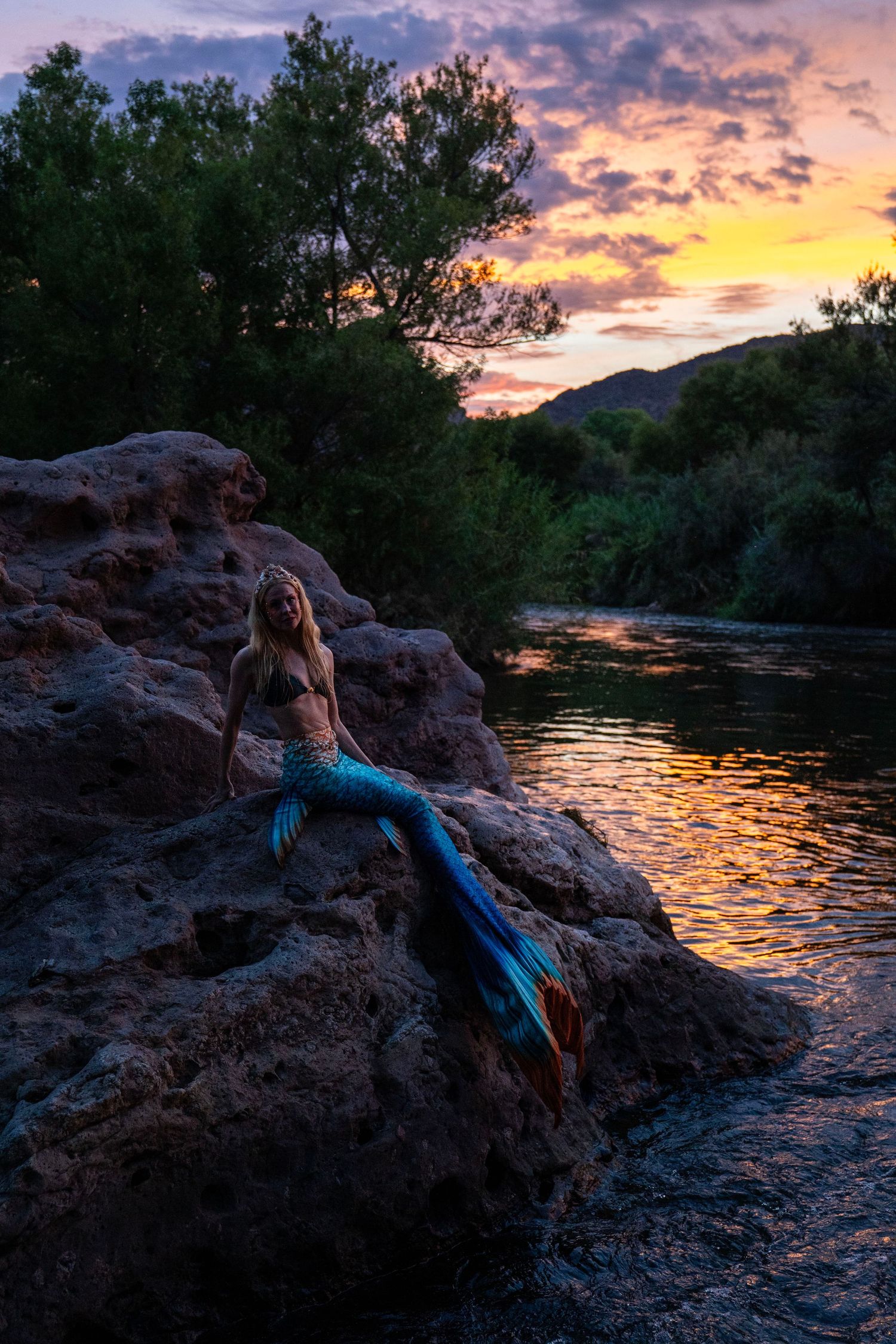 Mermaid cosplayer reclining on rocks beside a river during a vibrant sunset. Mermaid photoshoot, fantasy photoshoot. Arizona photographer.